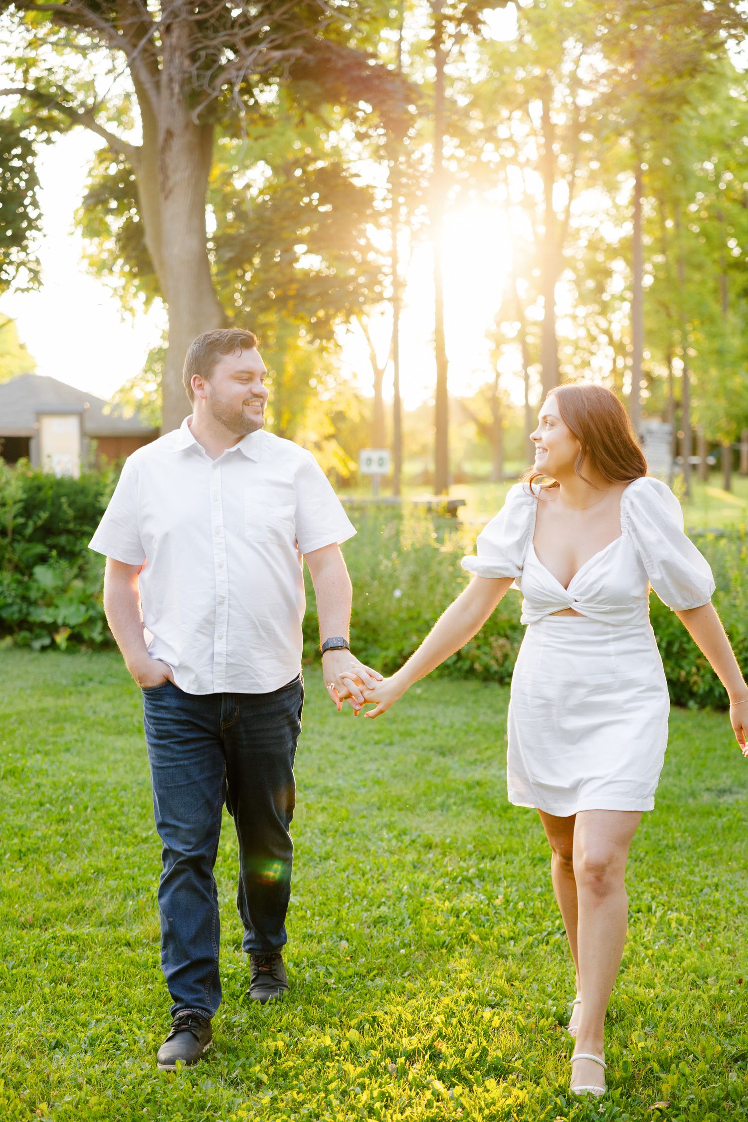 Couple holding hands while walking through sunlit garden in white summer outfits.