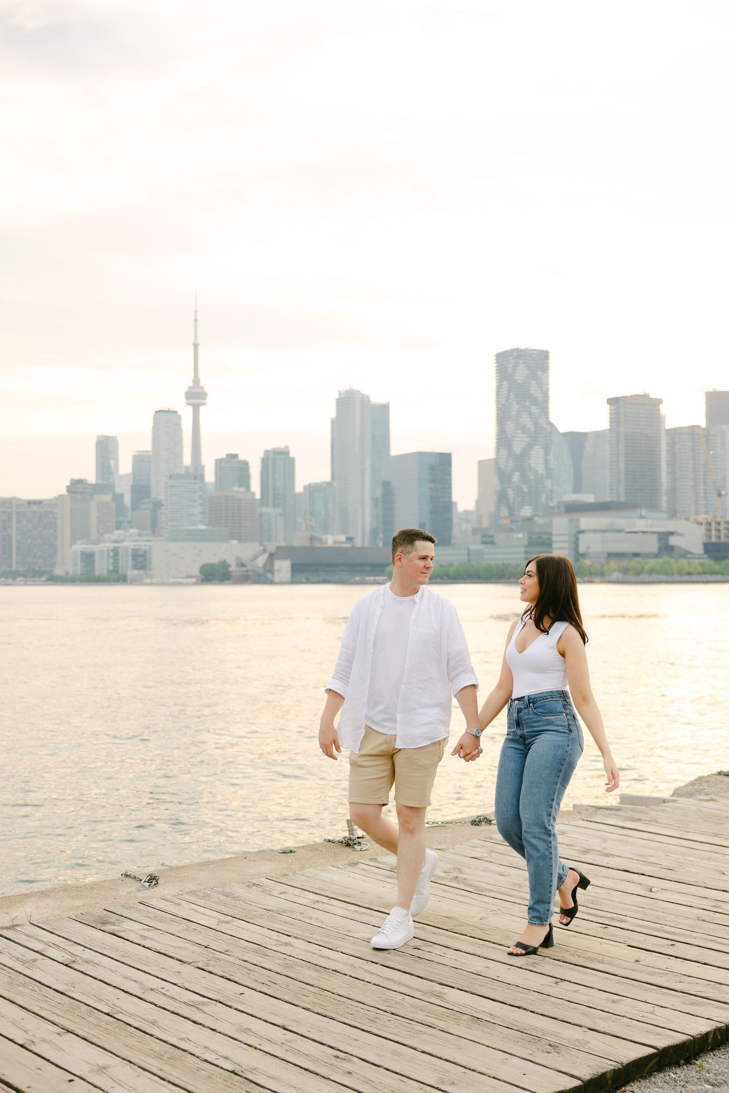 Couple walking hand in hand on a waterfront boardwalk with Toronto's CN Tower and cityscape behind them.