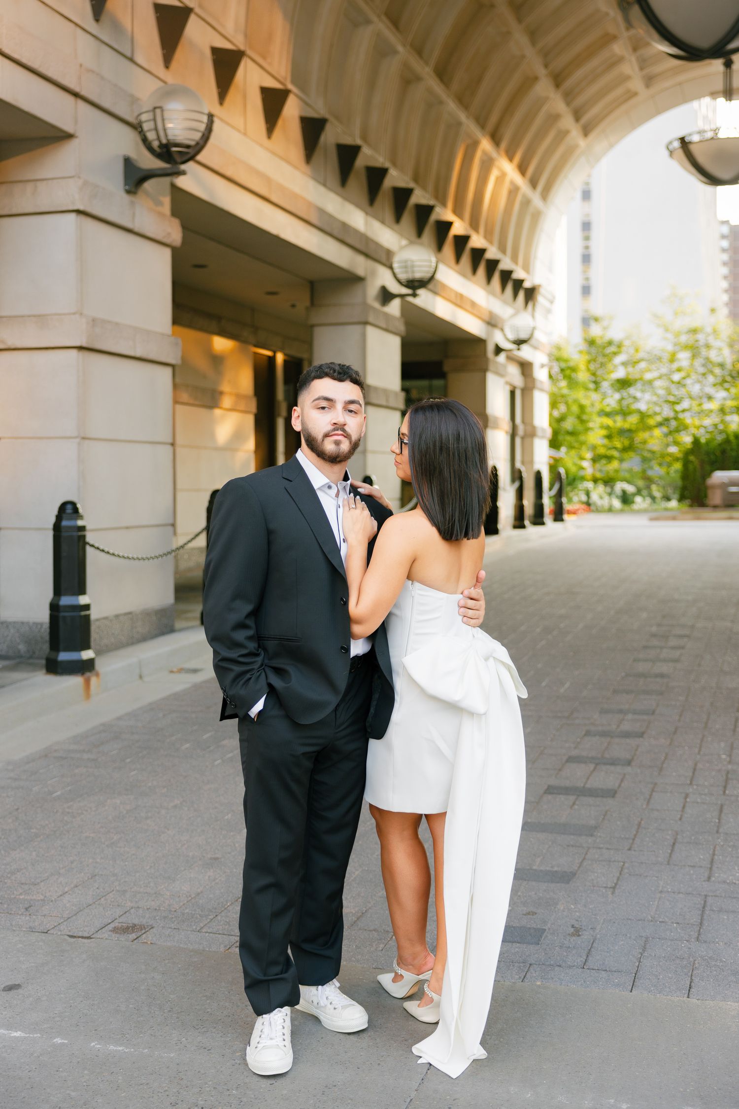 Elegantly dressed couple poses beneath architectural arches in an urban setting.