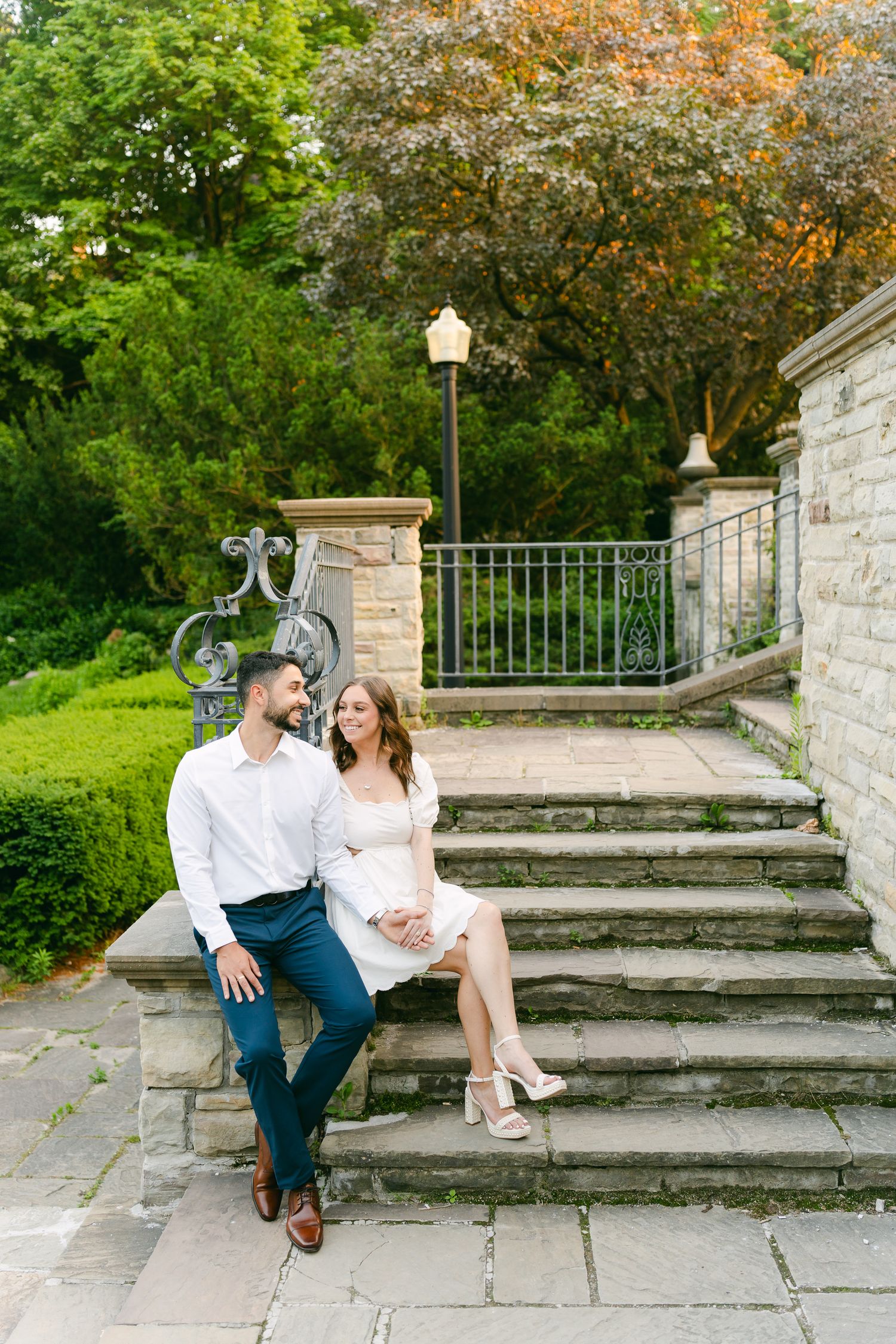 A relaxed portrait on garden stone steps with lush greenery and decorative lamp post in background.