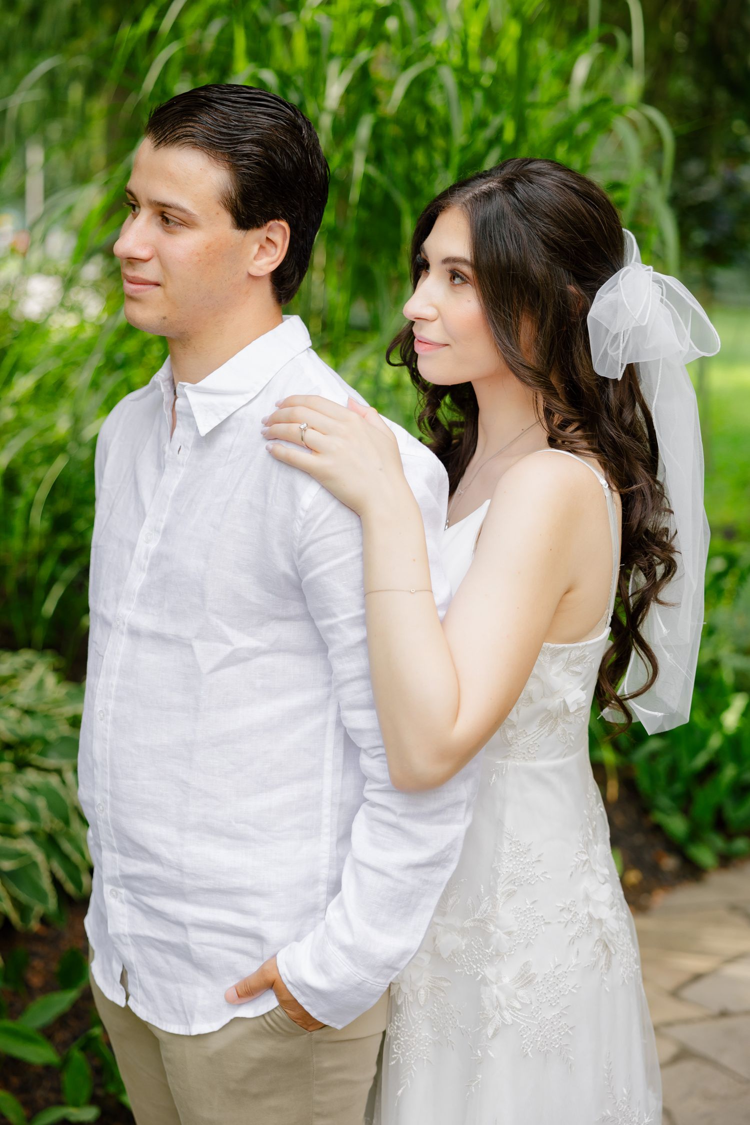 A wedding couple poses in a garden setting, wearing white attire with a lush green backdrop.