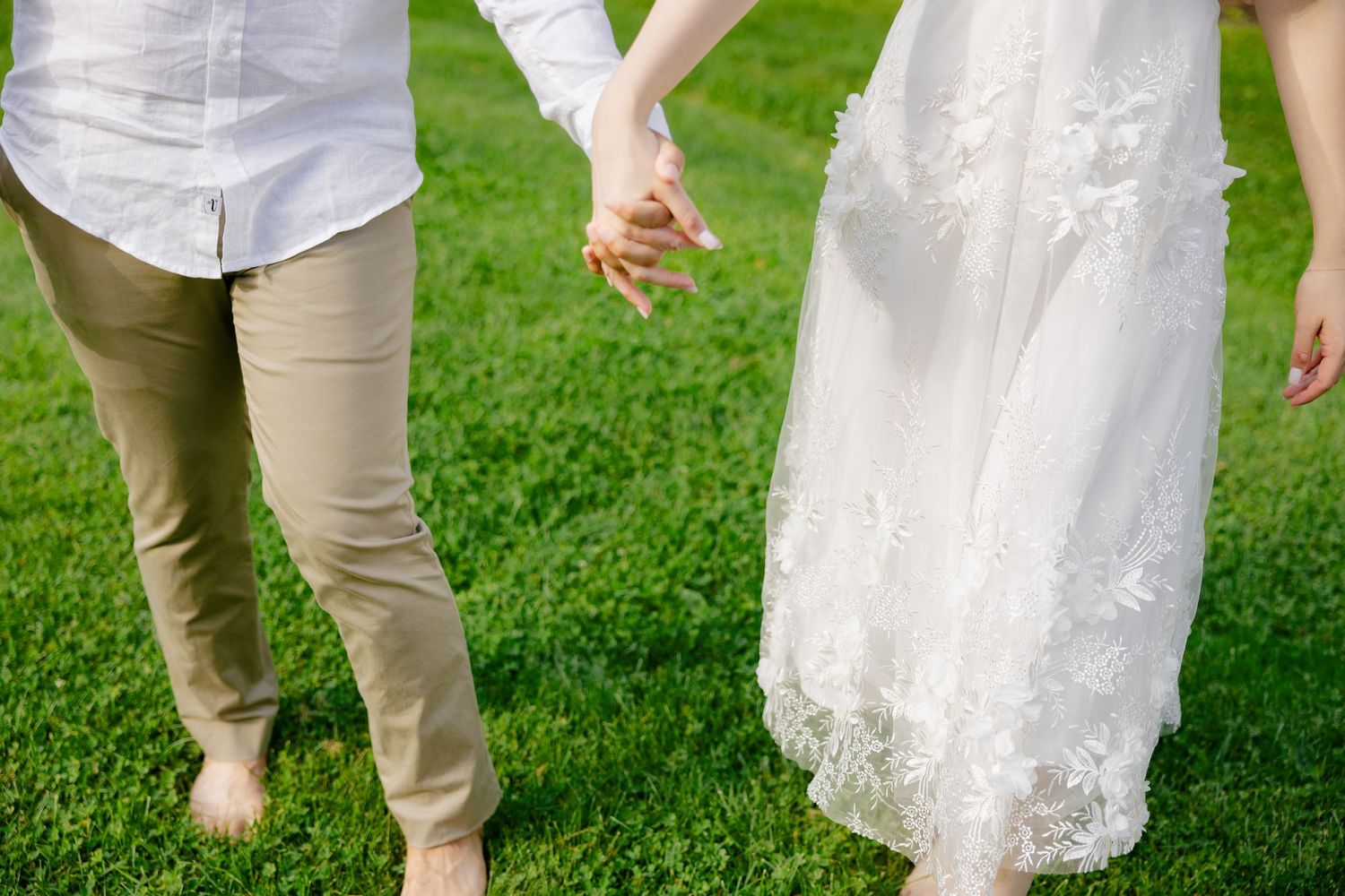 A couple holding hands barefoot on green grass, with the bride wearing a lace wedding dress and groom in casual attire.