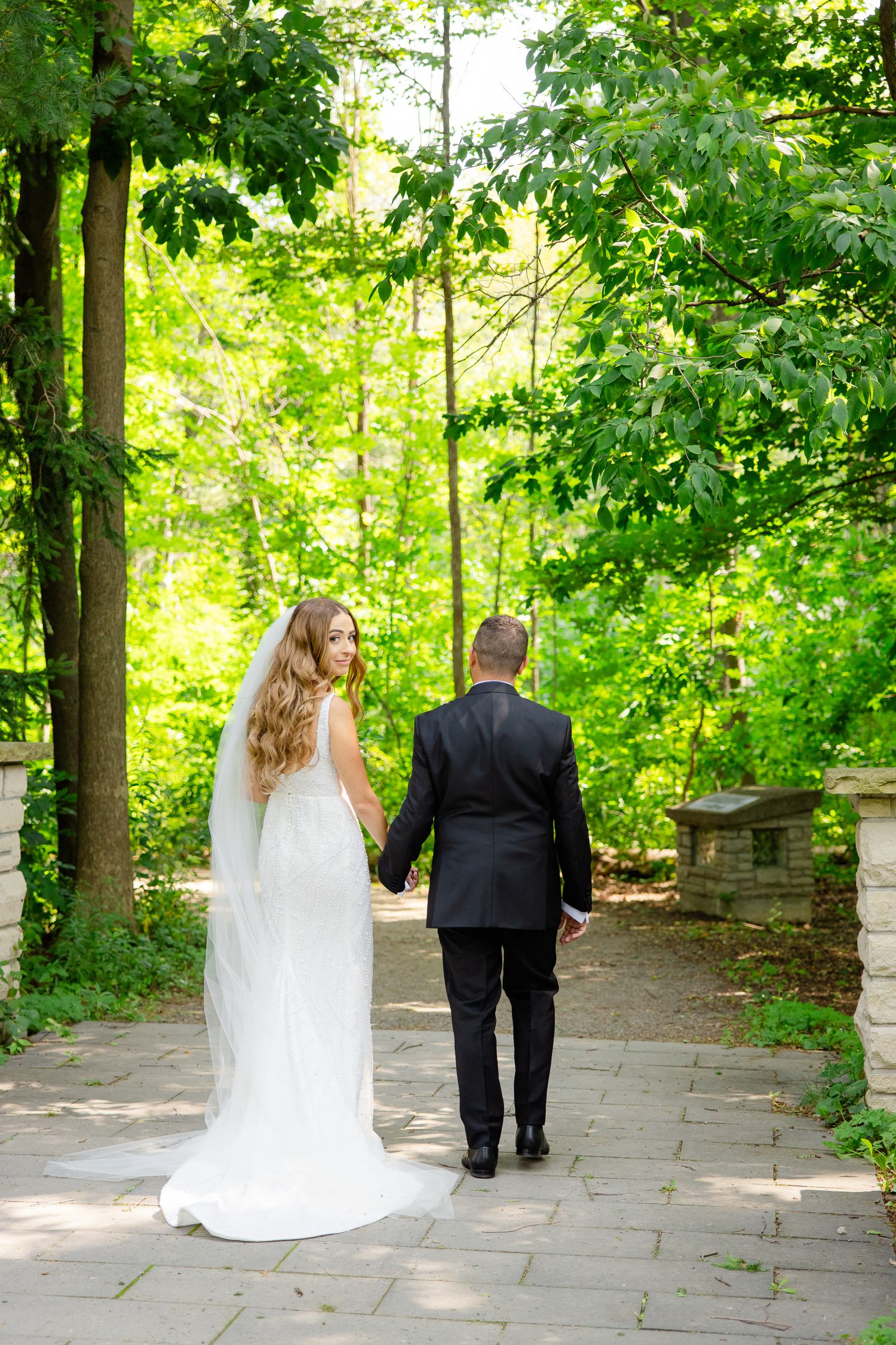 Newlyweds holding hands while walking down stone pathway surrounded by trees.