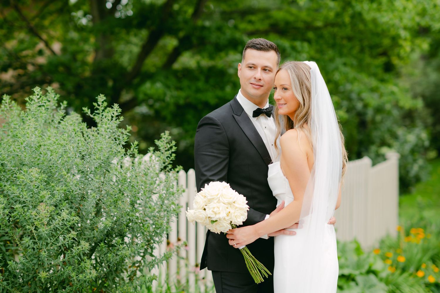 Wedding couple poses with white bouquet in front of lush garden setting and white fence.