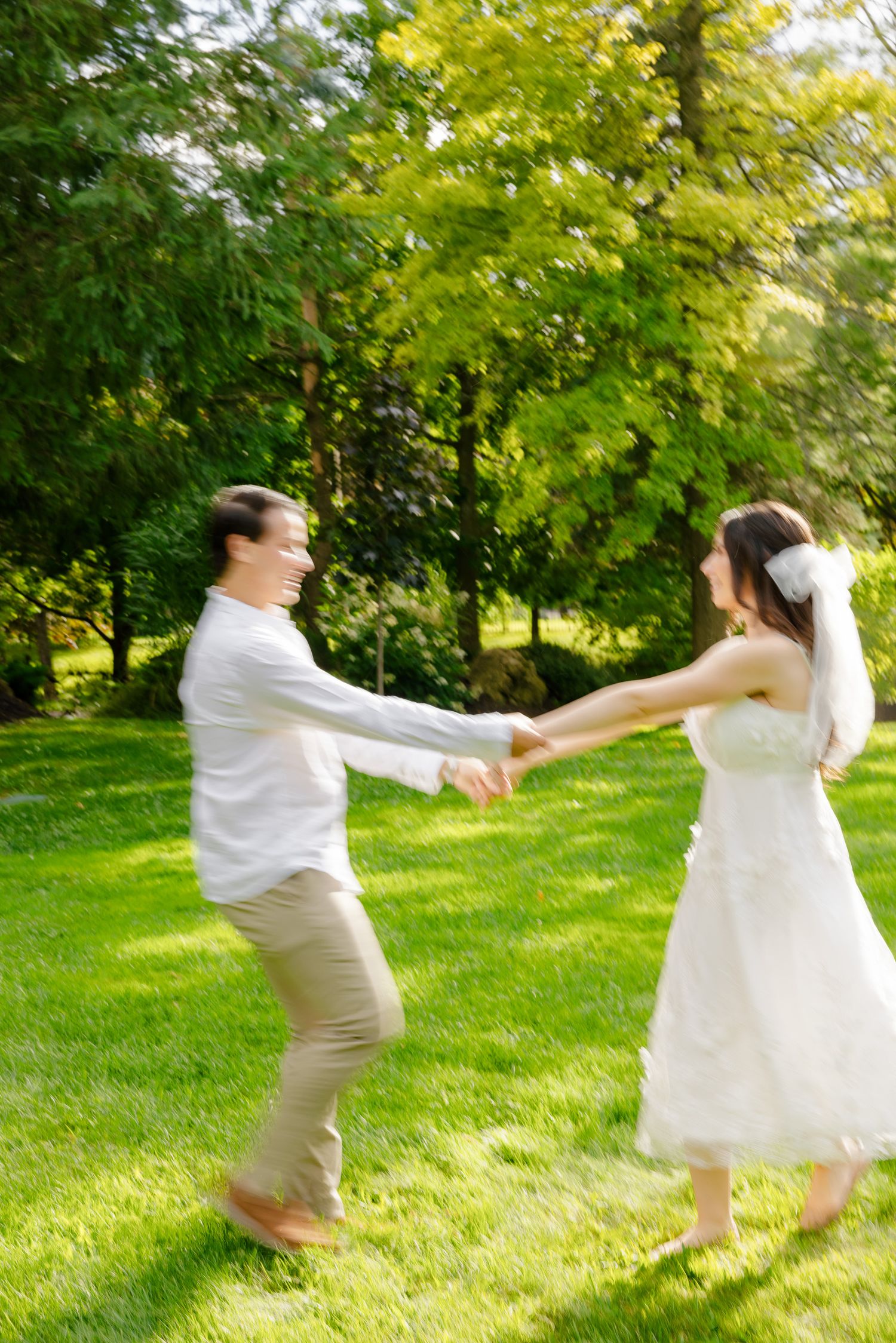 A couple in wedding attire dances and spins in a sunny garden with lush greenery.