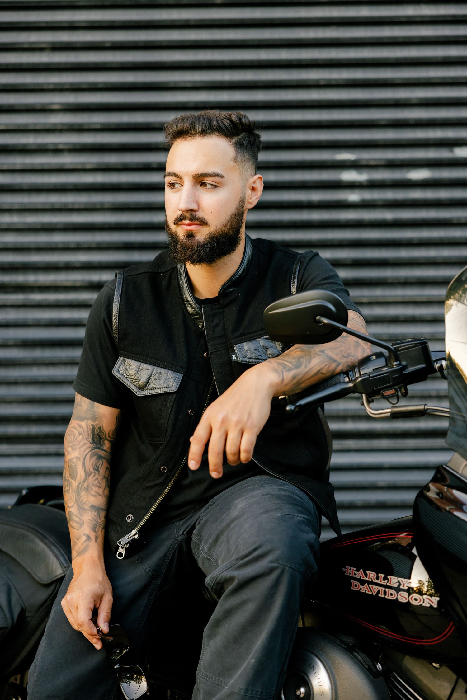 A tattooed biker in black clothing sits casually on a parked motorcycle against corrugated metal backdrop.
