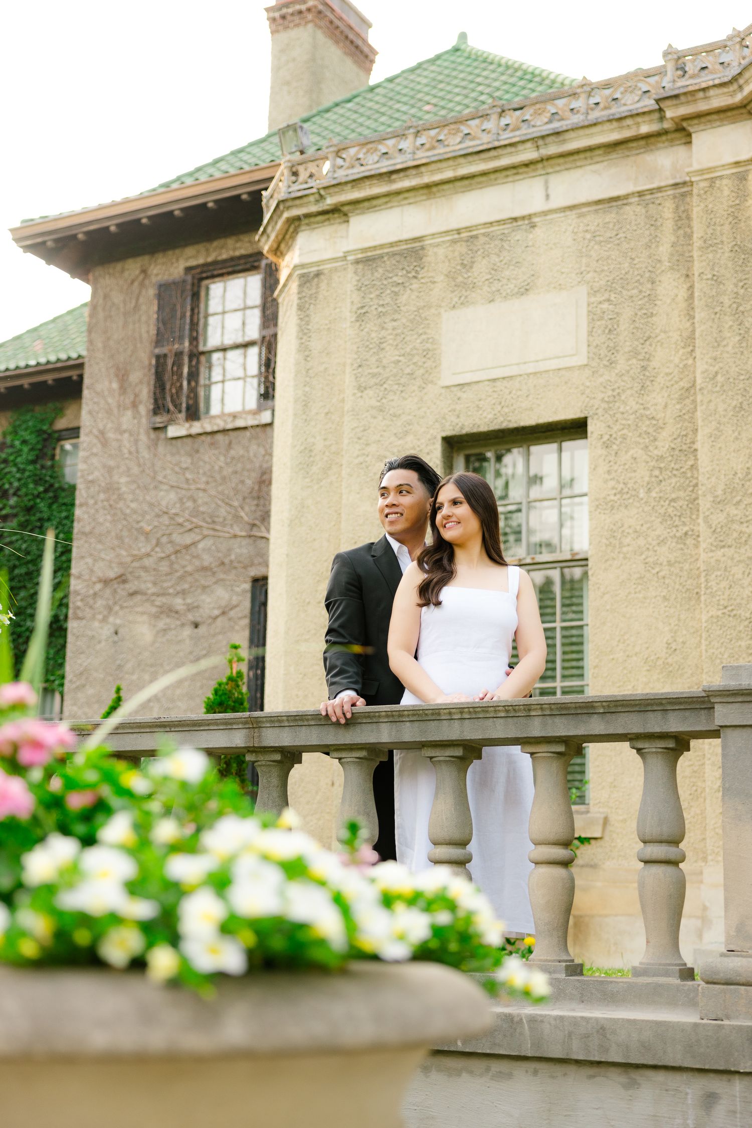 A couple stands at an elegant stone balustrade against a historic building with a green roof.