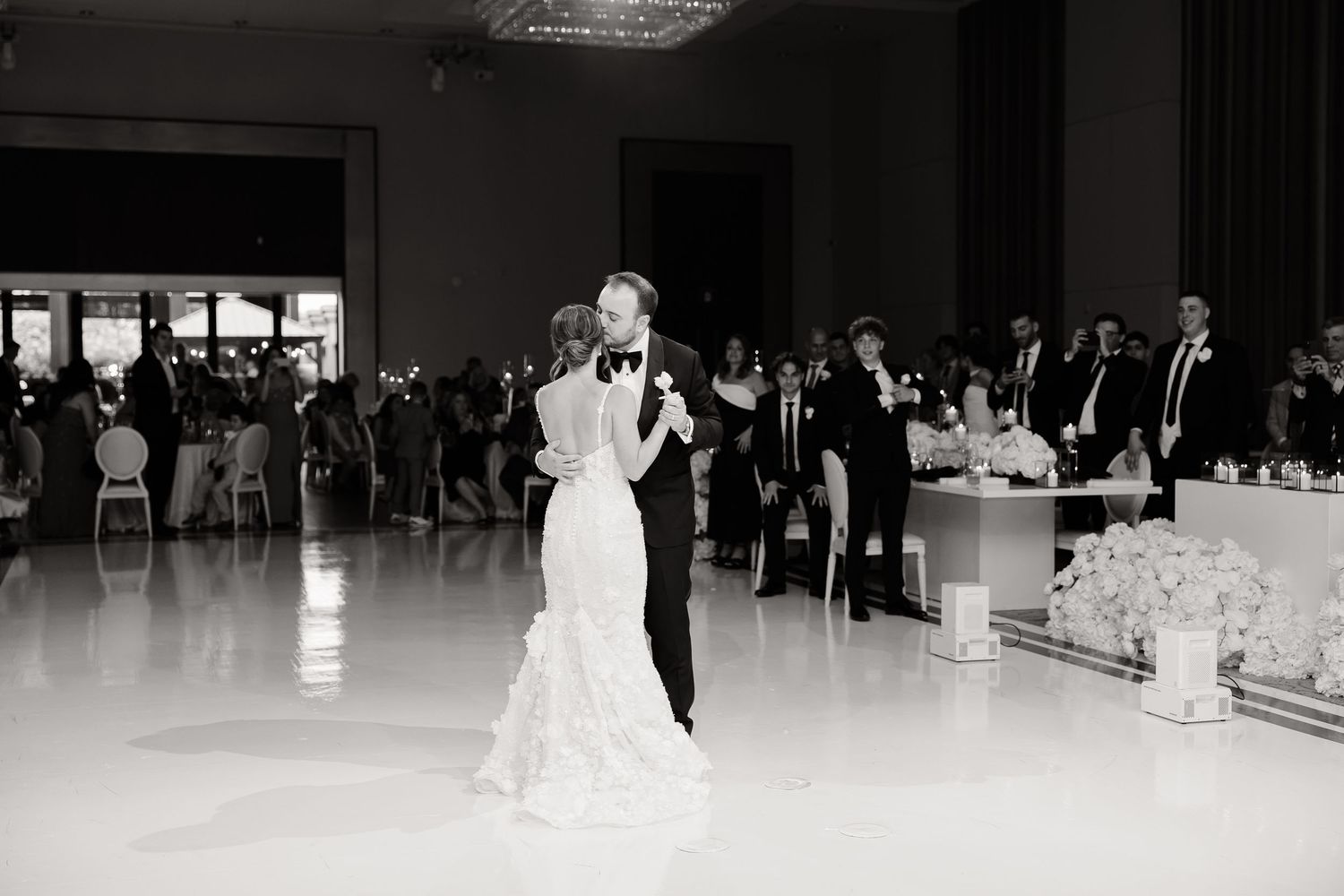 A black and white photograph of a couple sharing their first dance at their wedding reception in an elegant ballroom.