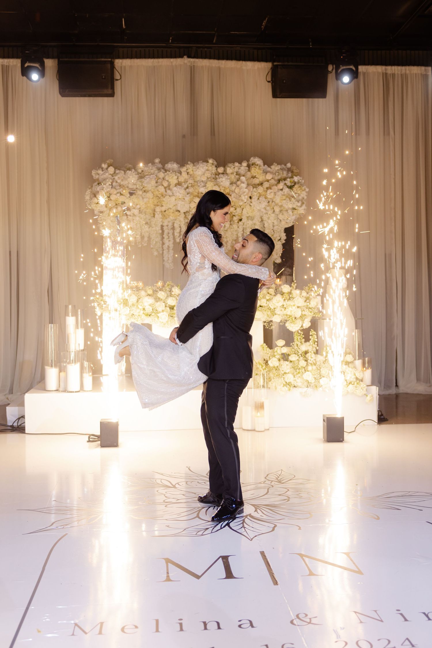 Dynamic wedding dance lift with sparkler effects under floral arch on monogrammed floor.