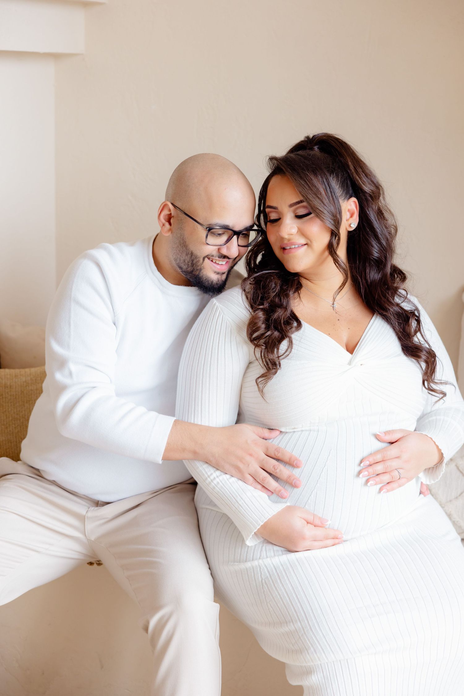 Expecting couple in matching white outfits share intimate moment on couch during maternity photoshoot.