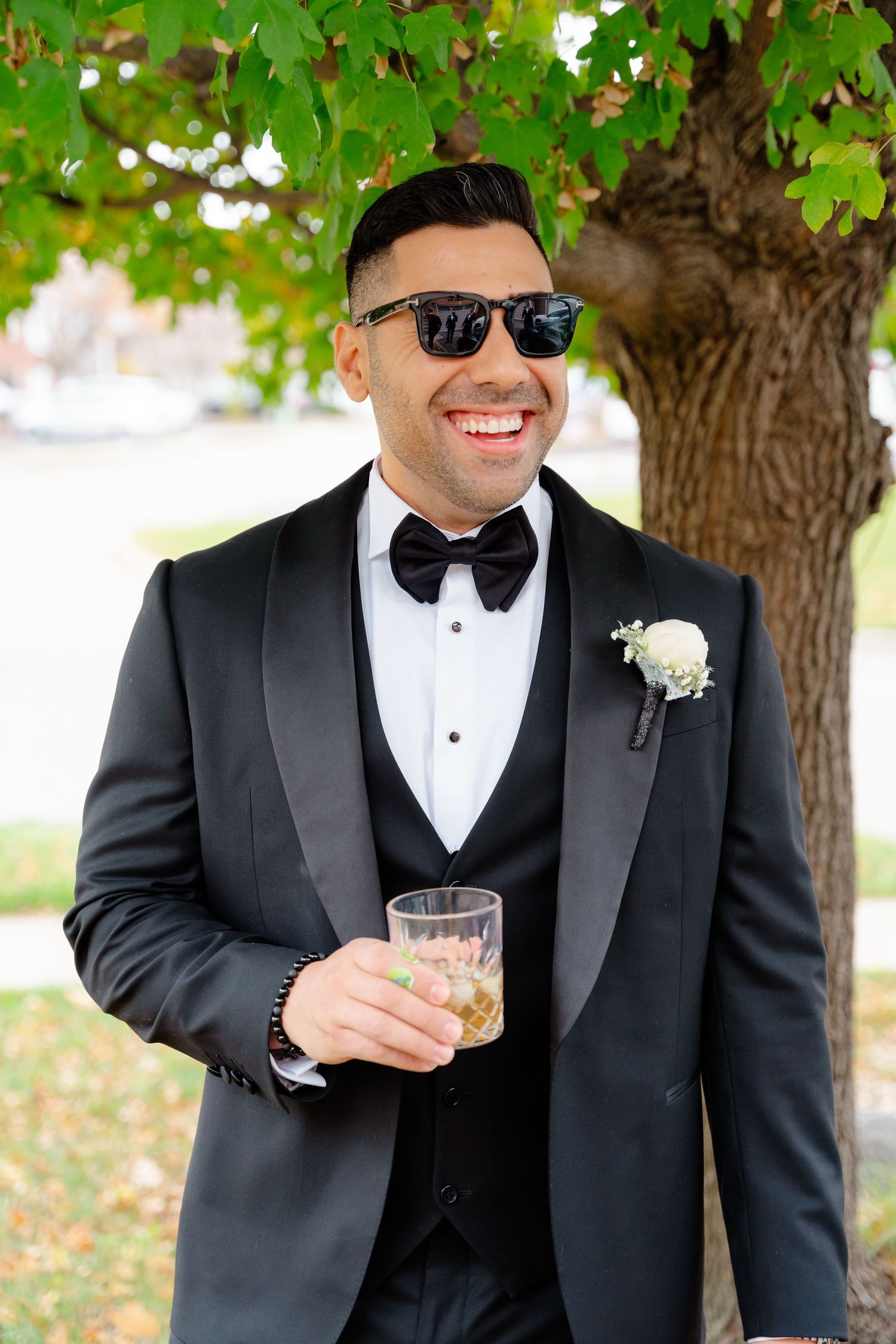 Wedding party member in black tuxedo smiling while holding drink under green leaves.