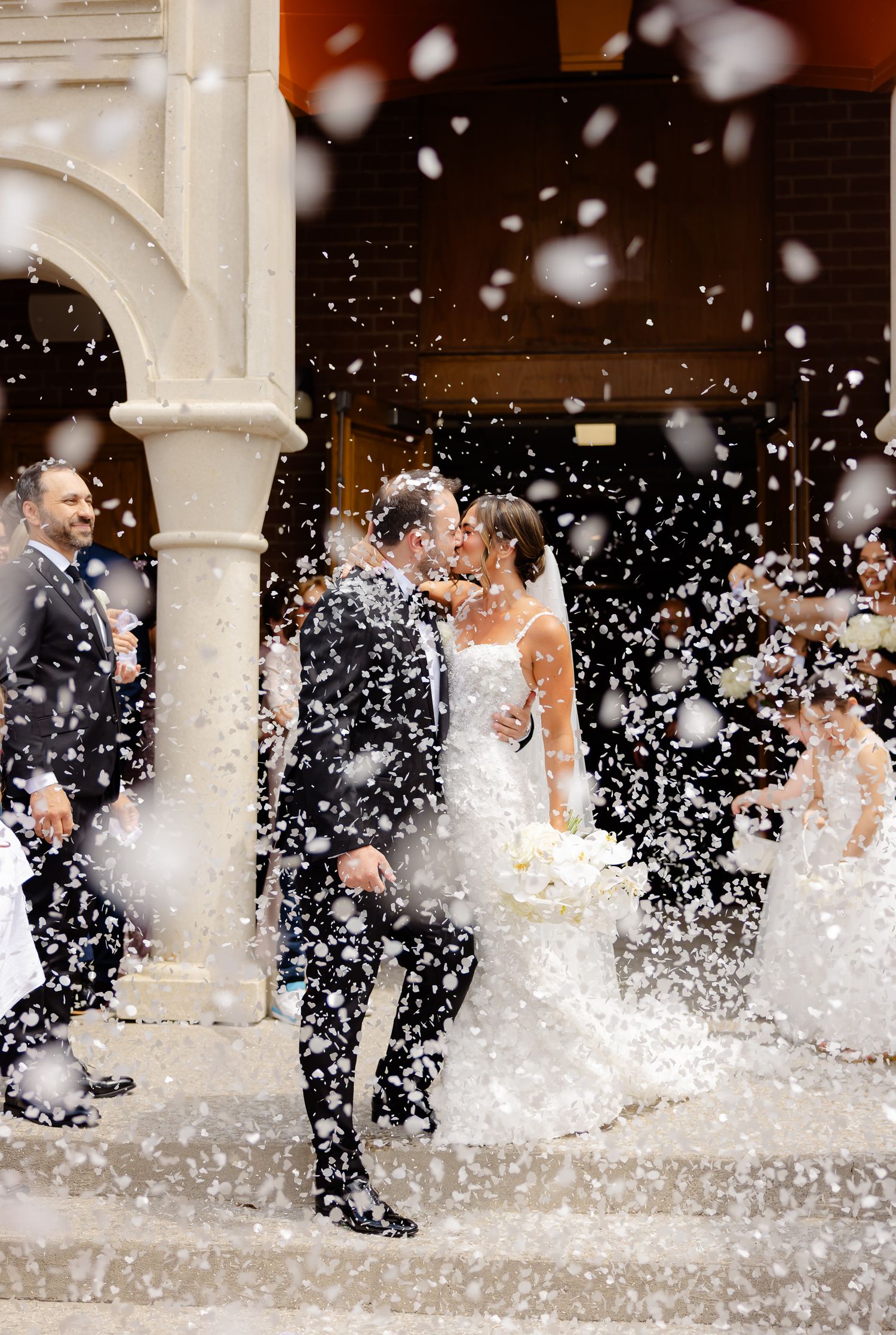 Wedding celebration with guests and falling confetti petals outside a grand architectural entrance with white columns.