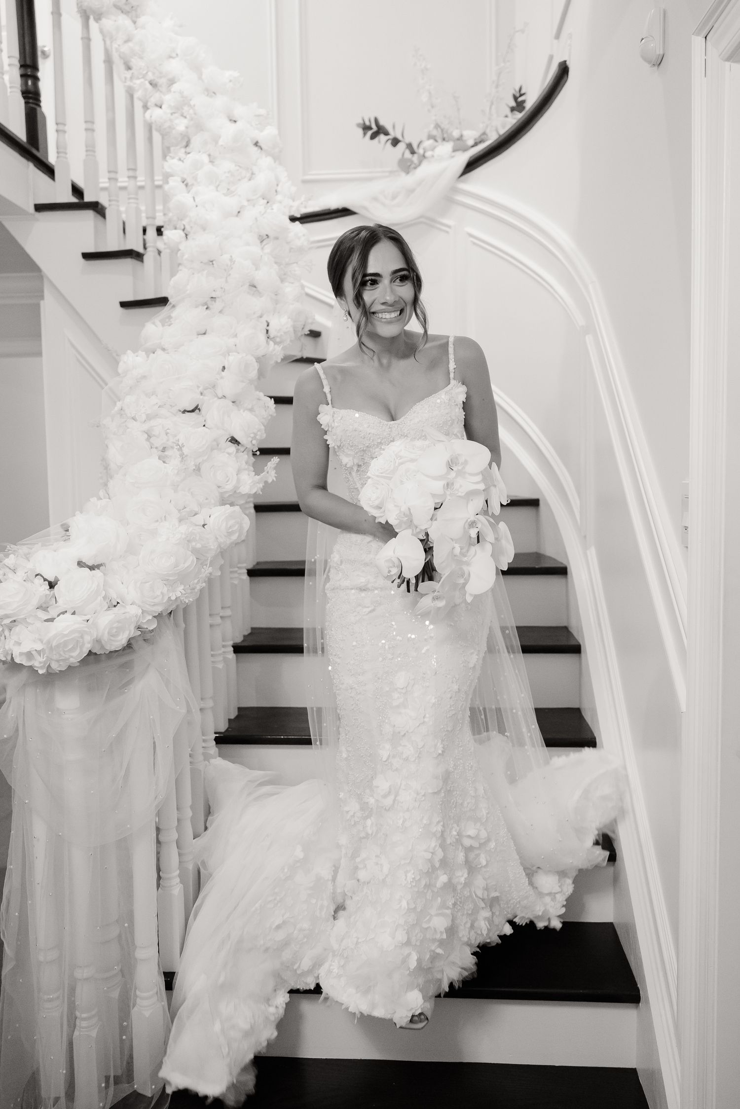 A bride in a lace wedding gown descends an elegant staircase decorated with white floral arrangements and tulle.