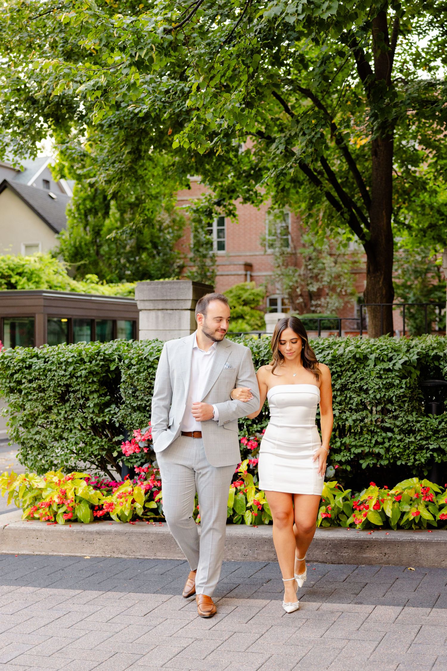 A stylish couple walks together on a brick path surrounded by lush gardens and trees.