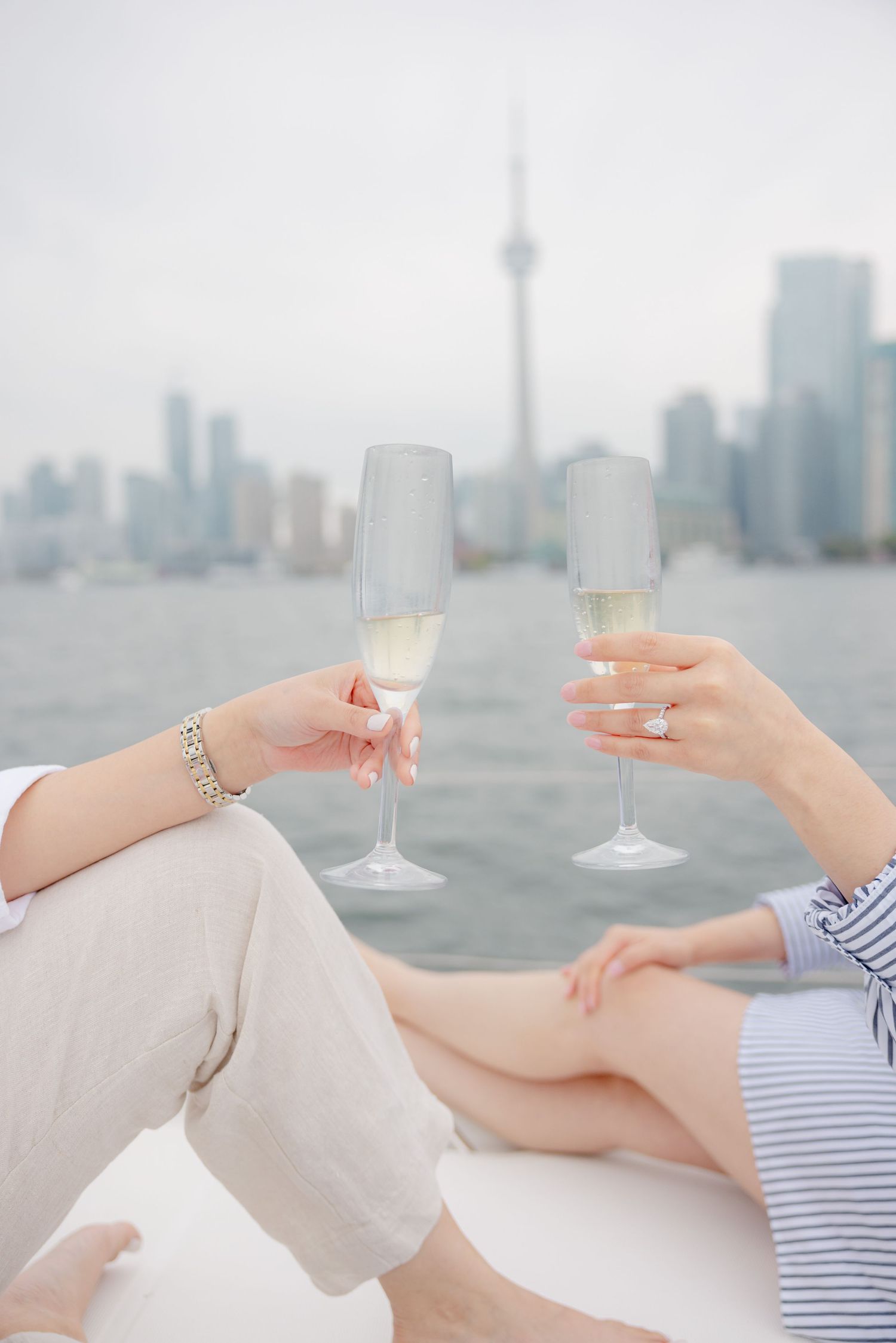 Two champagne glasses clink with Toronto's CN Tower skyline visible across the harbor on a misty day.