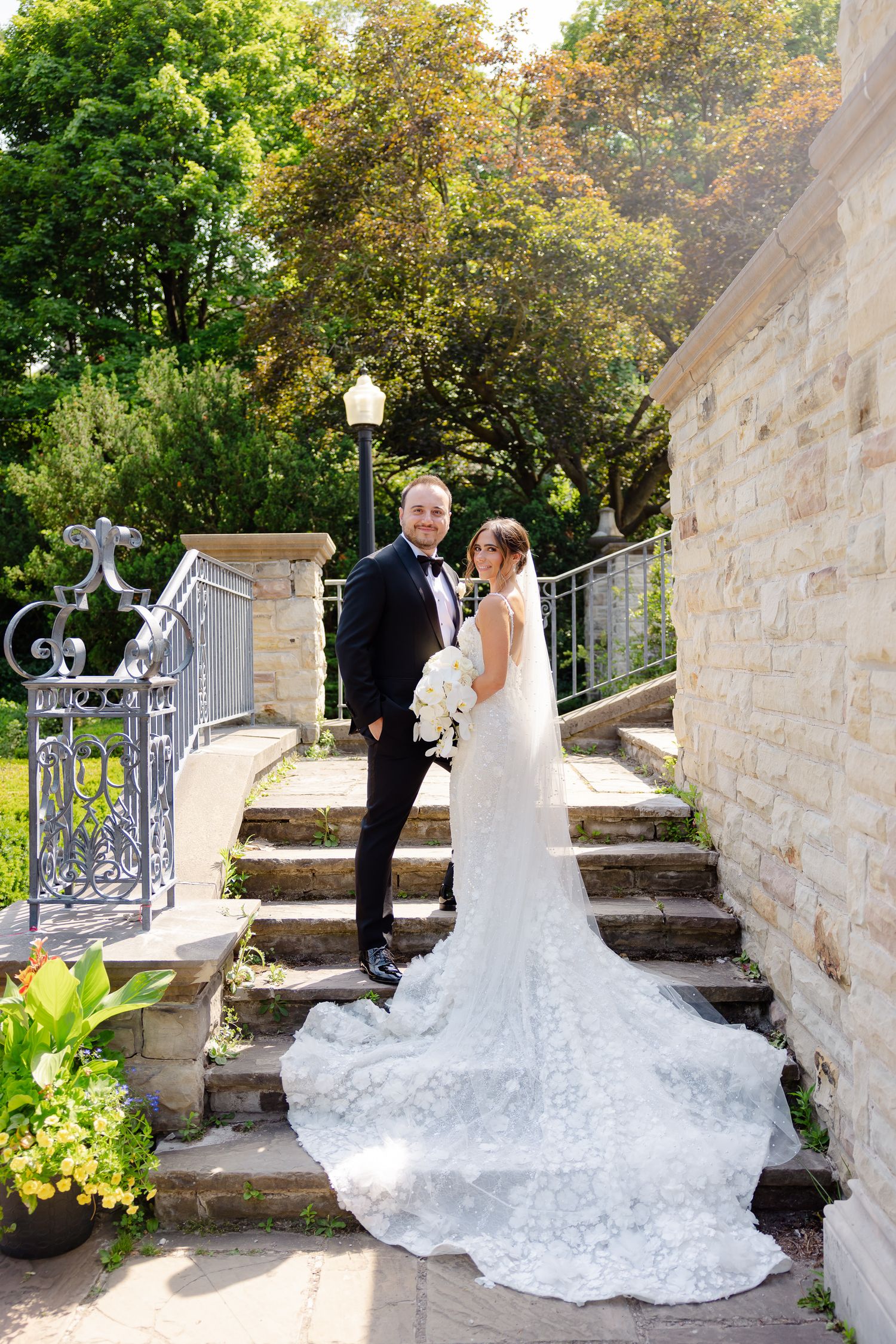 A couple in formal wedding attire stands on stone steps with an ornate railing and lush greenery backdrop.