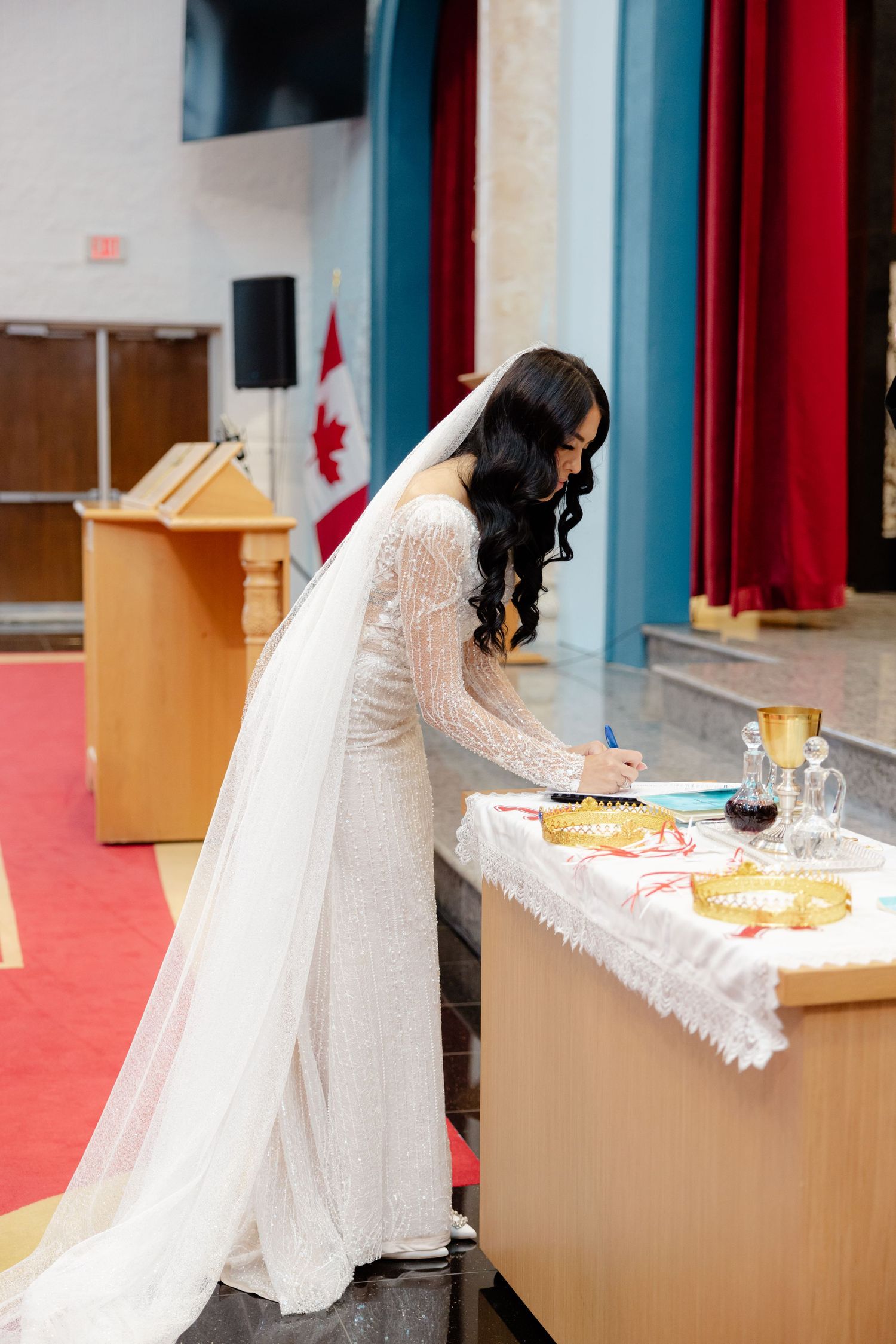A bride in a long white dress signs documents at a table during her wedding ceremony.
