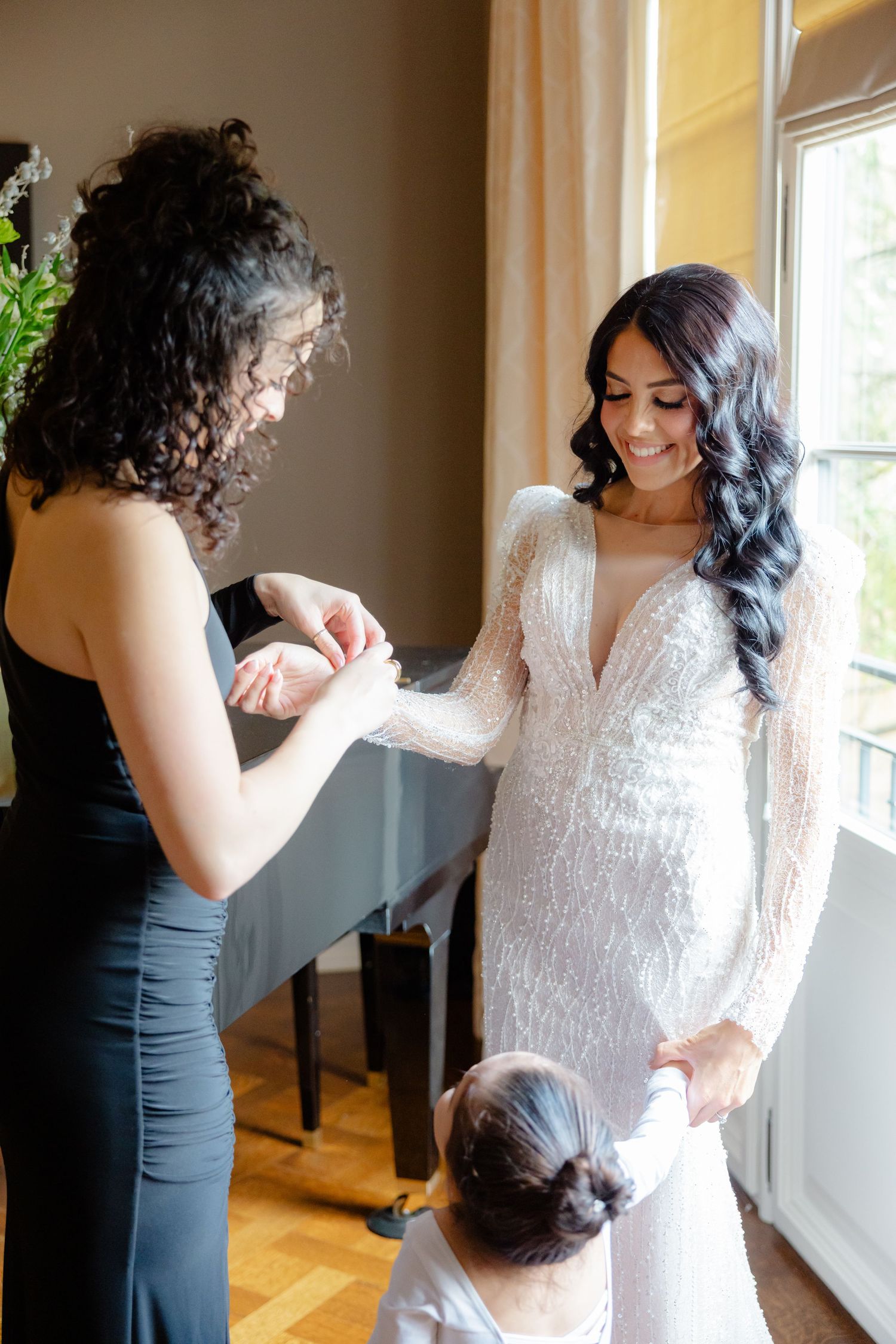 Long-sleeved lace wedding dress being fitted in sunny room with wood floors.