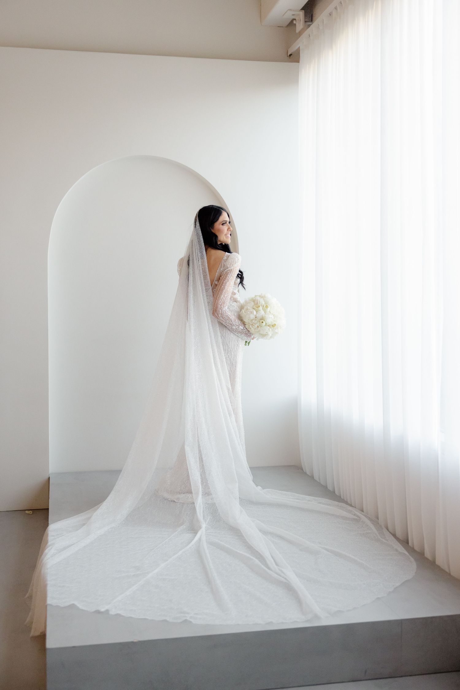A bride in a flowing white veil holds a bouquet while posing in an elegant white archway.