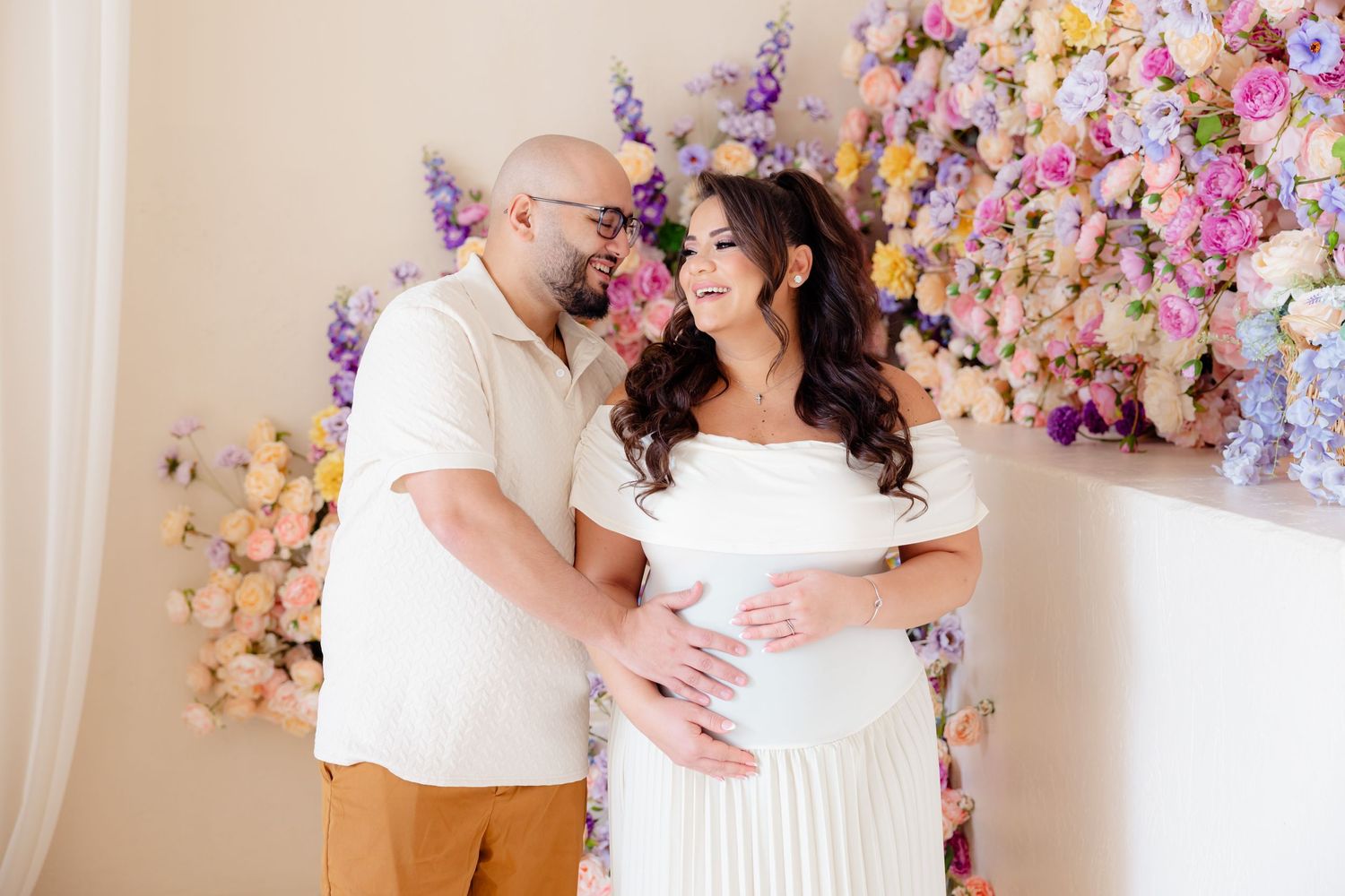 A couple poses together against a wall decorated with colorful spring flowers during a maternity photoshoot.