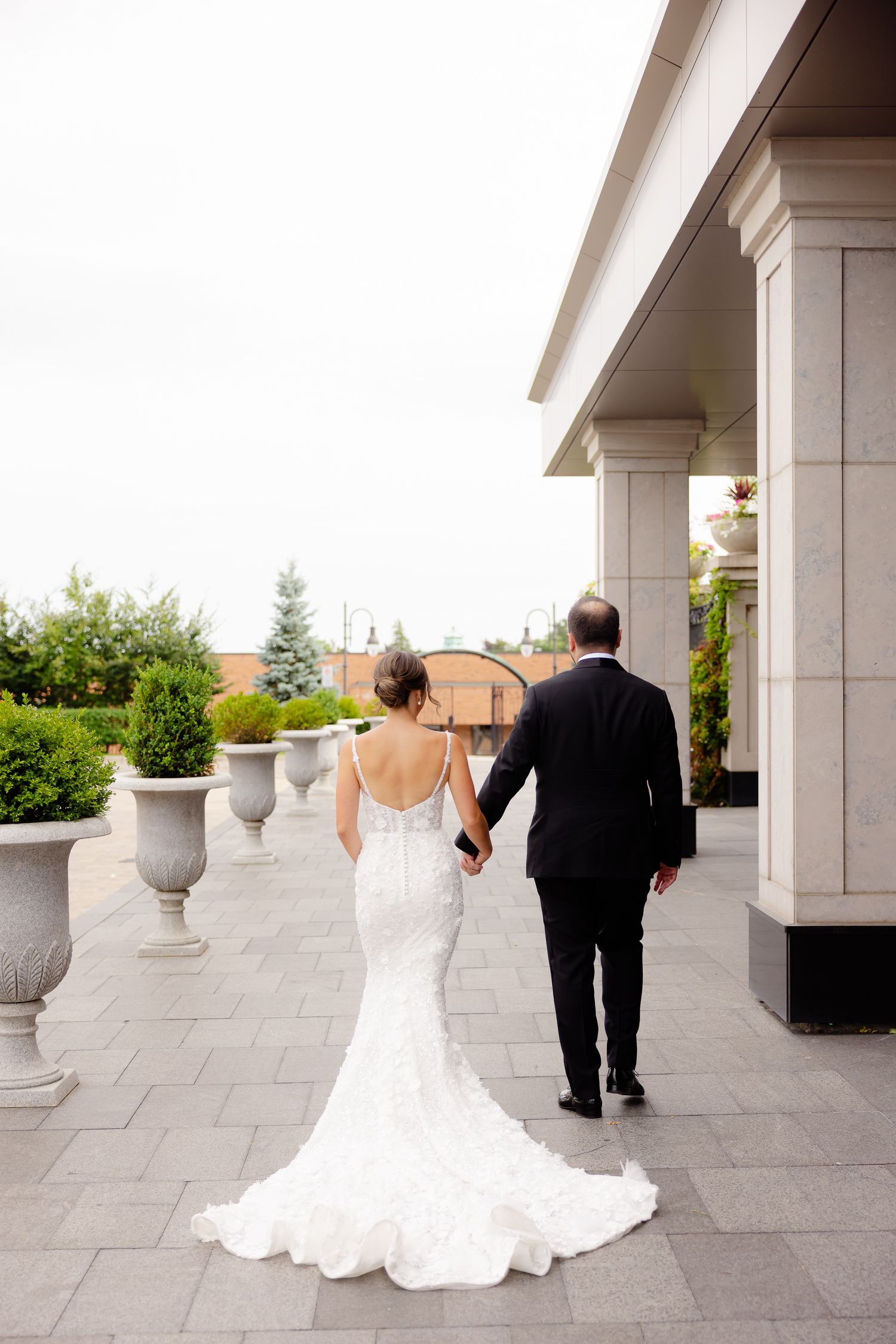 A couple in wedding attire walks along an elegant stone terrace with decorative urns and columns on a sunny day.