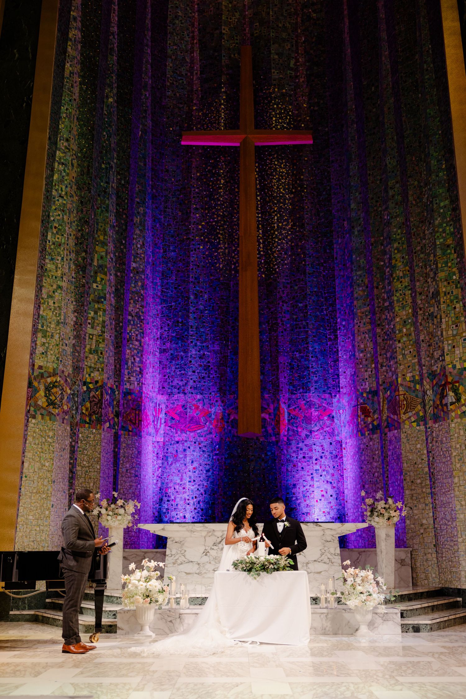 Wedding ceremony under illuminated cross with purple lighting and white floral decorations.