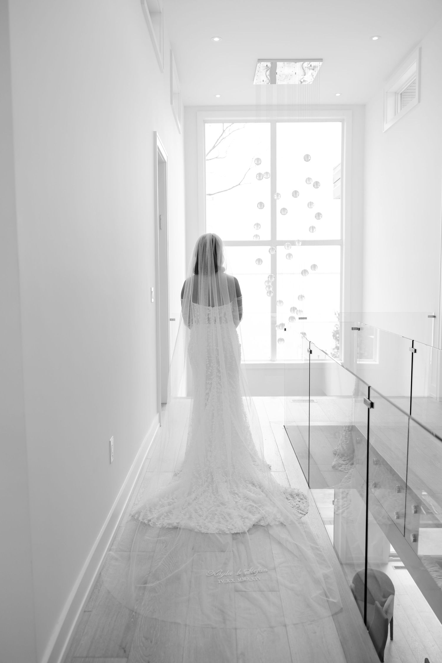 Back view of bride in lace gown with cathedral veil standing in bright white hallway.