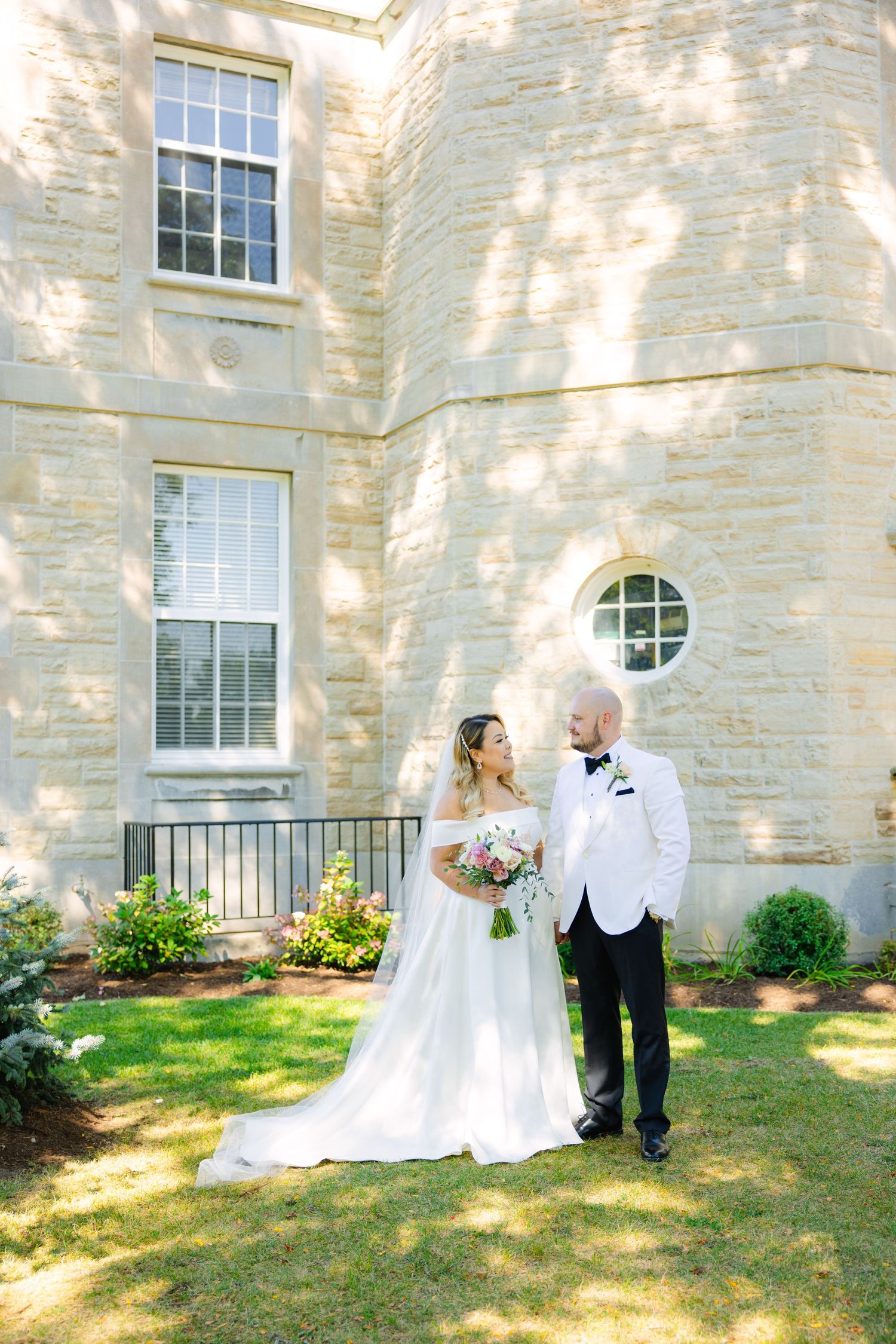 A romantic wedding photo taken outside a historic stone building with dappled sunlight.