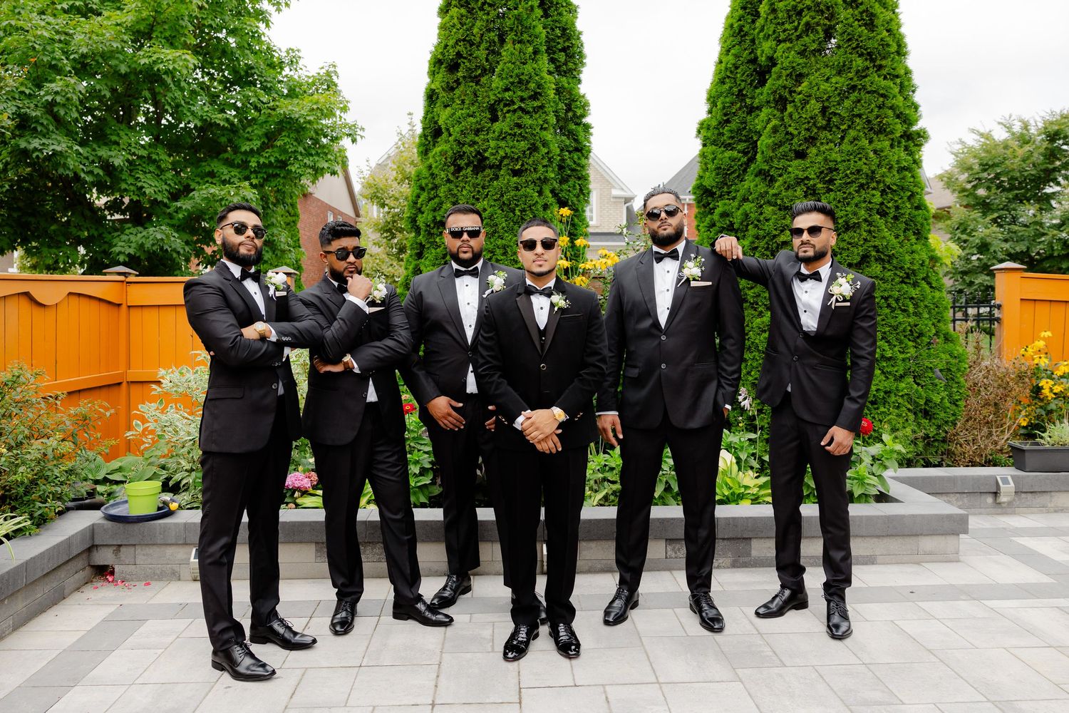 Wedding party in black tuxedos stands in formation against an orange fence and green cypress trees.