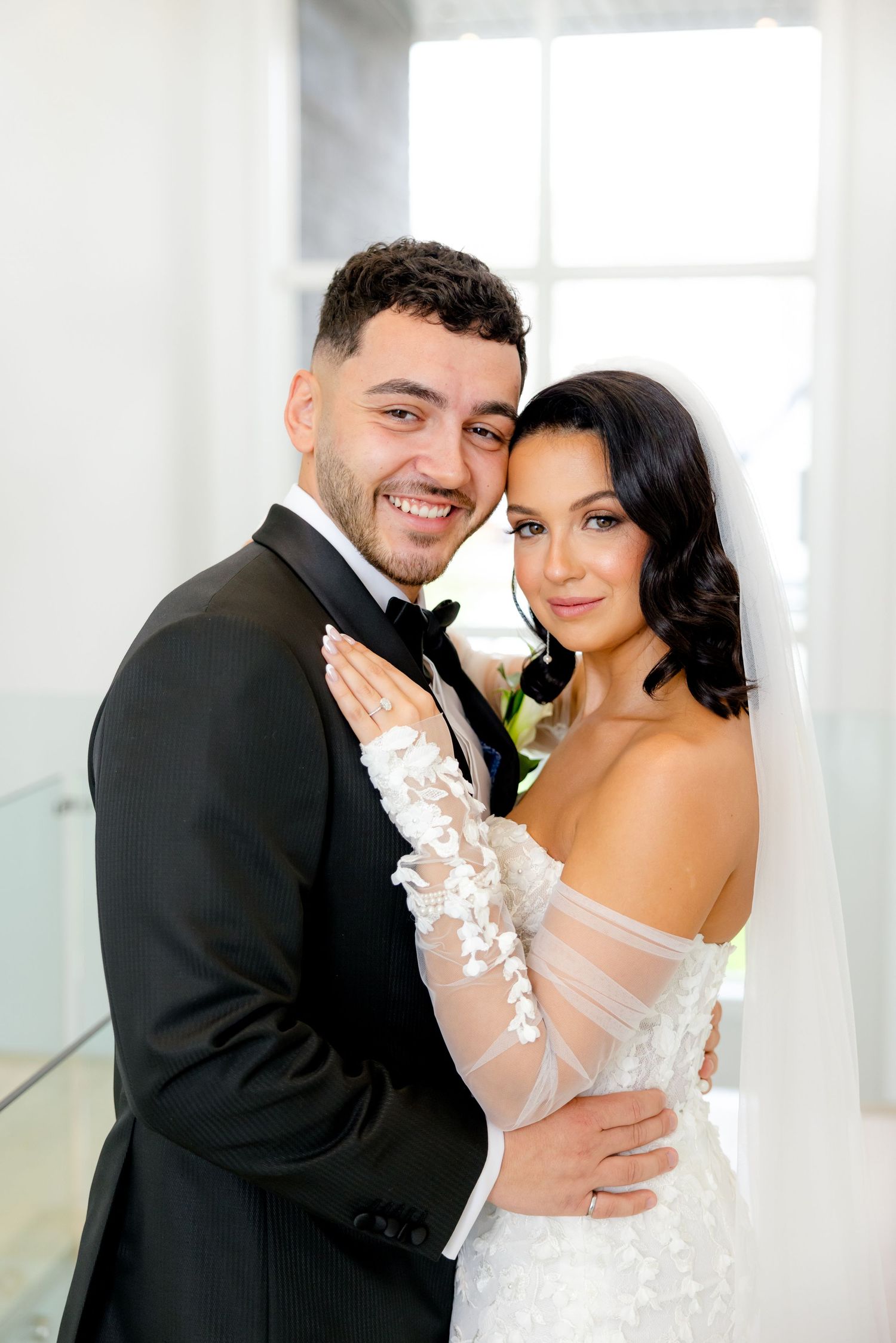 Elegant bride in lace off-shoulder gown embraces groom in classic black tuxedo during wedding portraits.