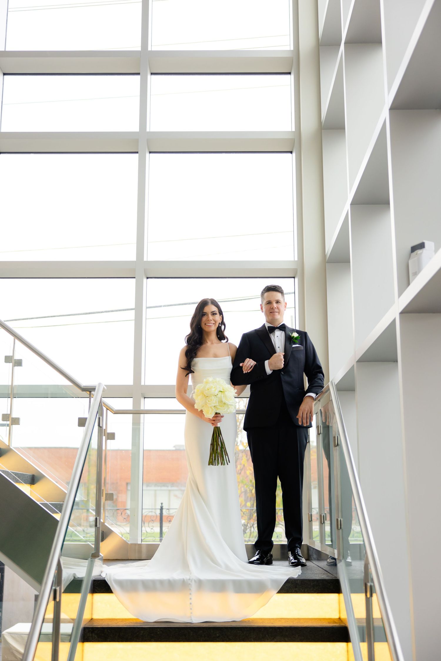Couple stands together on yellow-trimmed stairs in modern building with glass walls.
