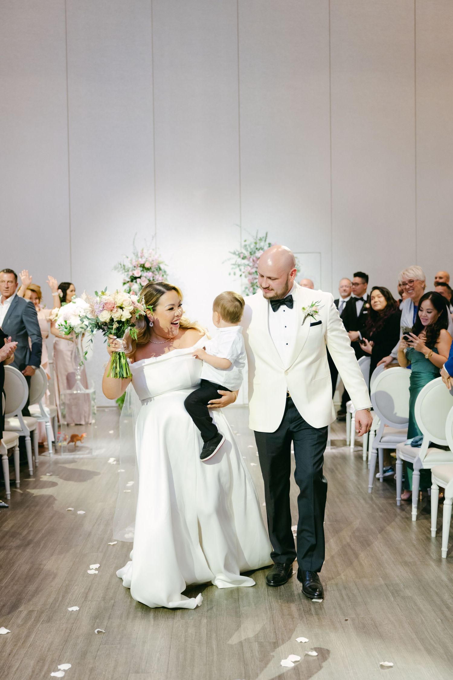 A joyful wedding recessional with a couple and small child walking down a petal-strewn aisle.