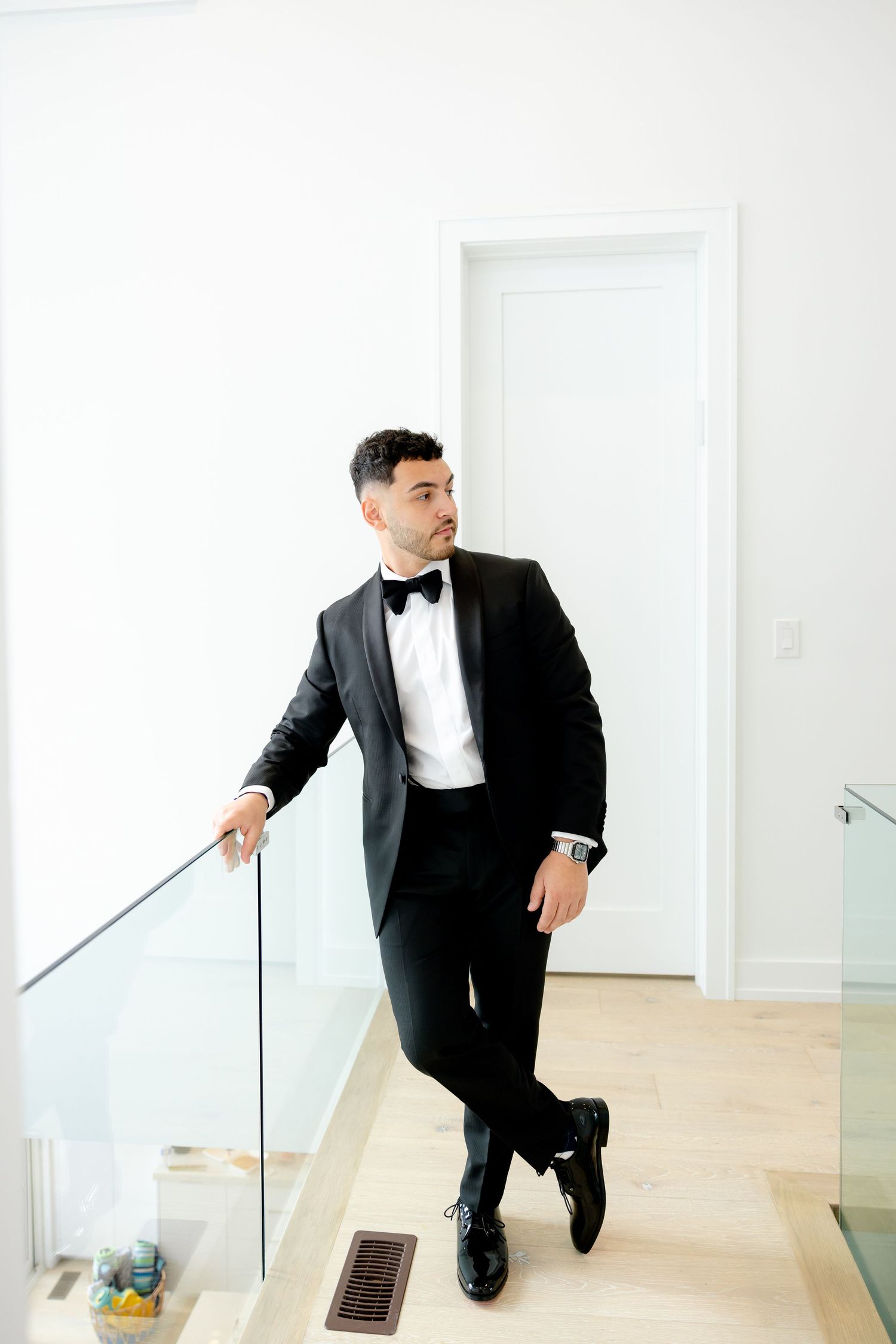 Groom in classic black tuxedo poses elegantly against white railing in modern interior setting.