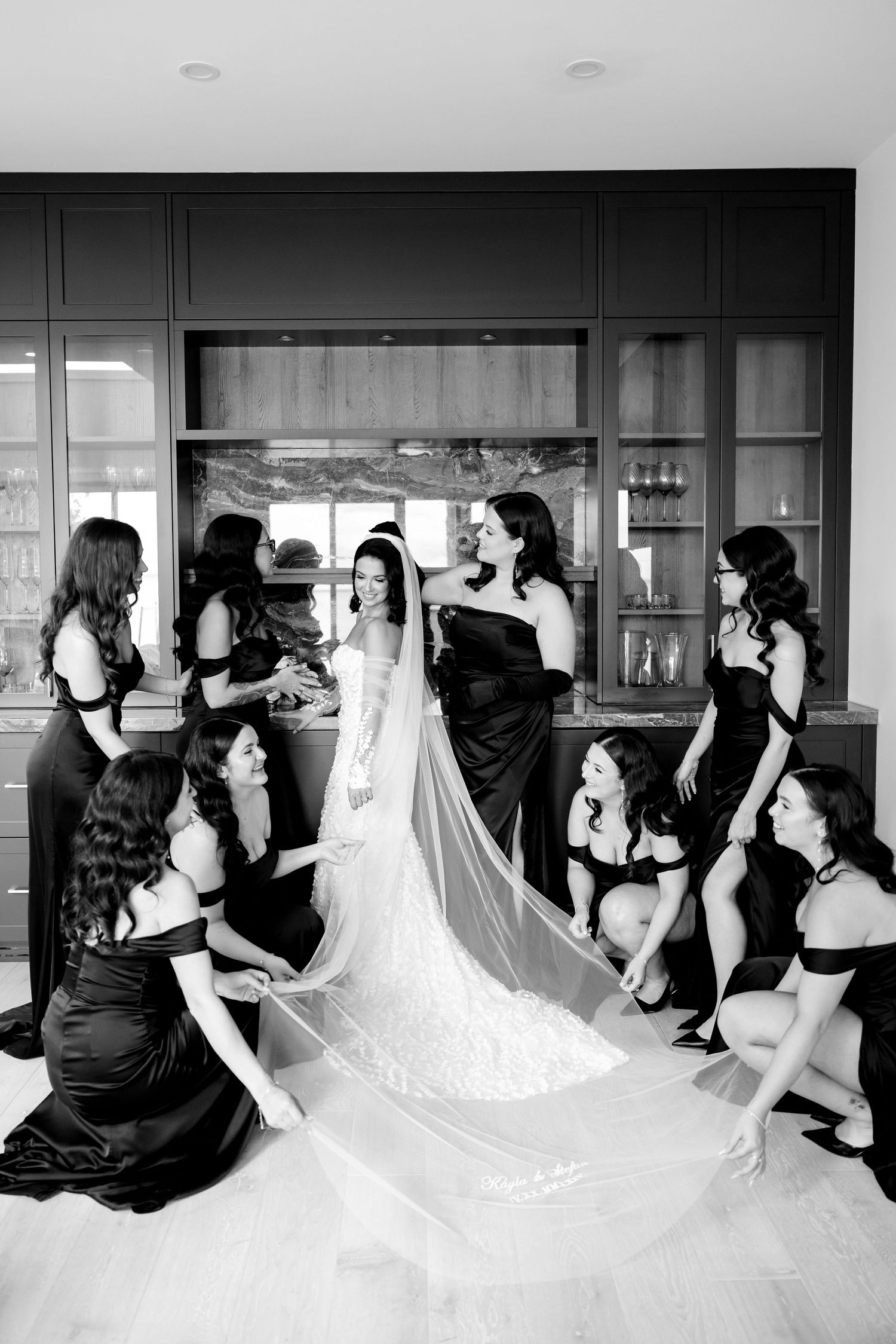 Black and white photo of bridesmaids arranging bride's veil in modern kitchen setting.
