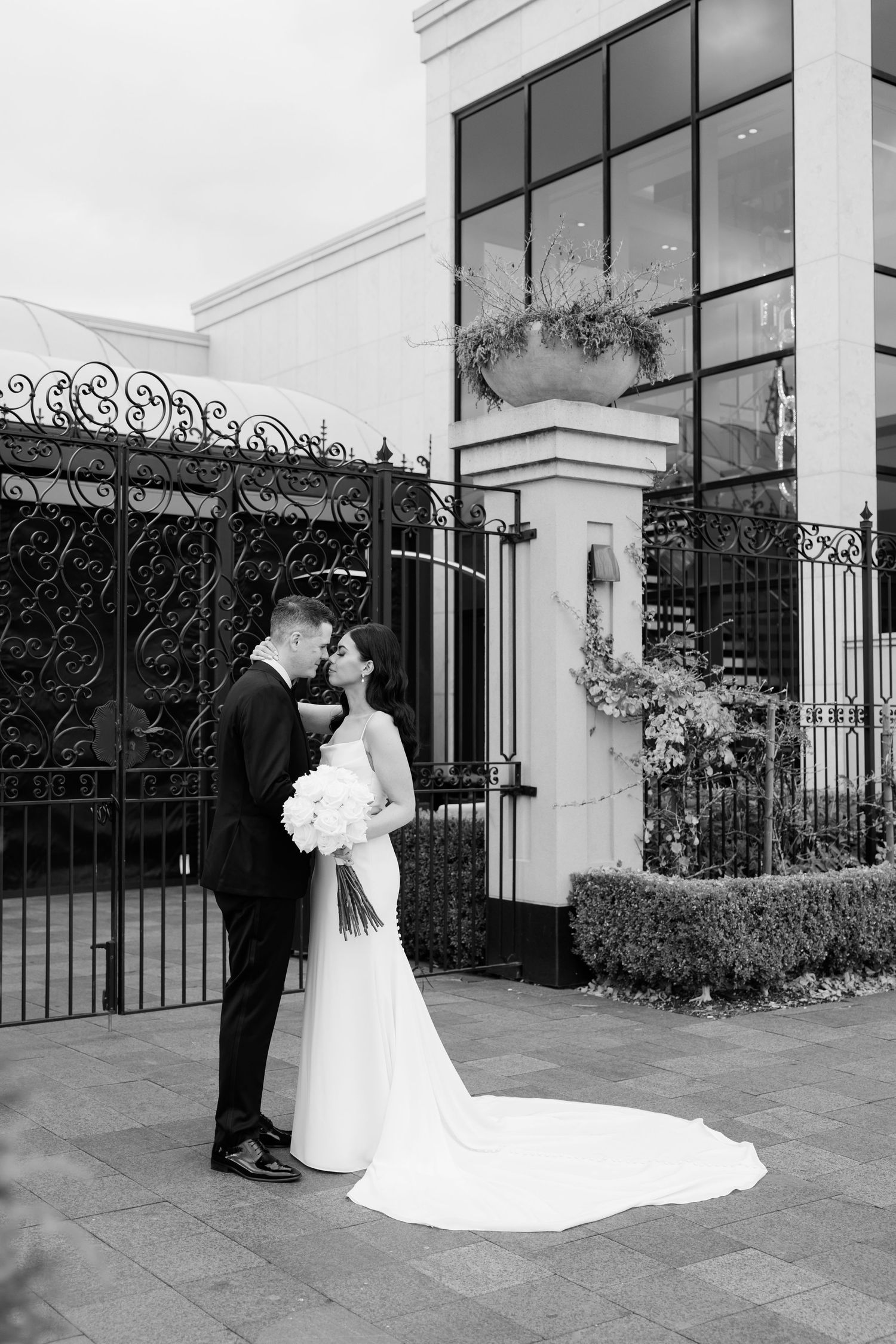 Black and white wedding photo showing couple embracing by architectural gates.