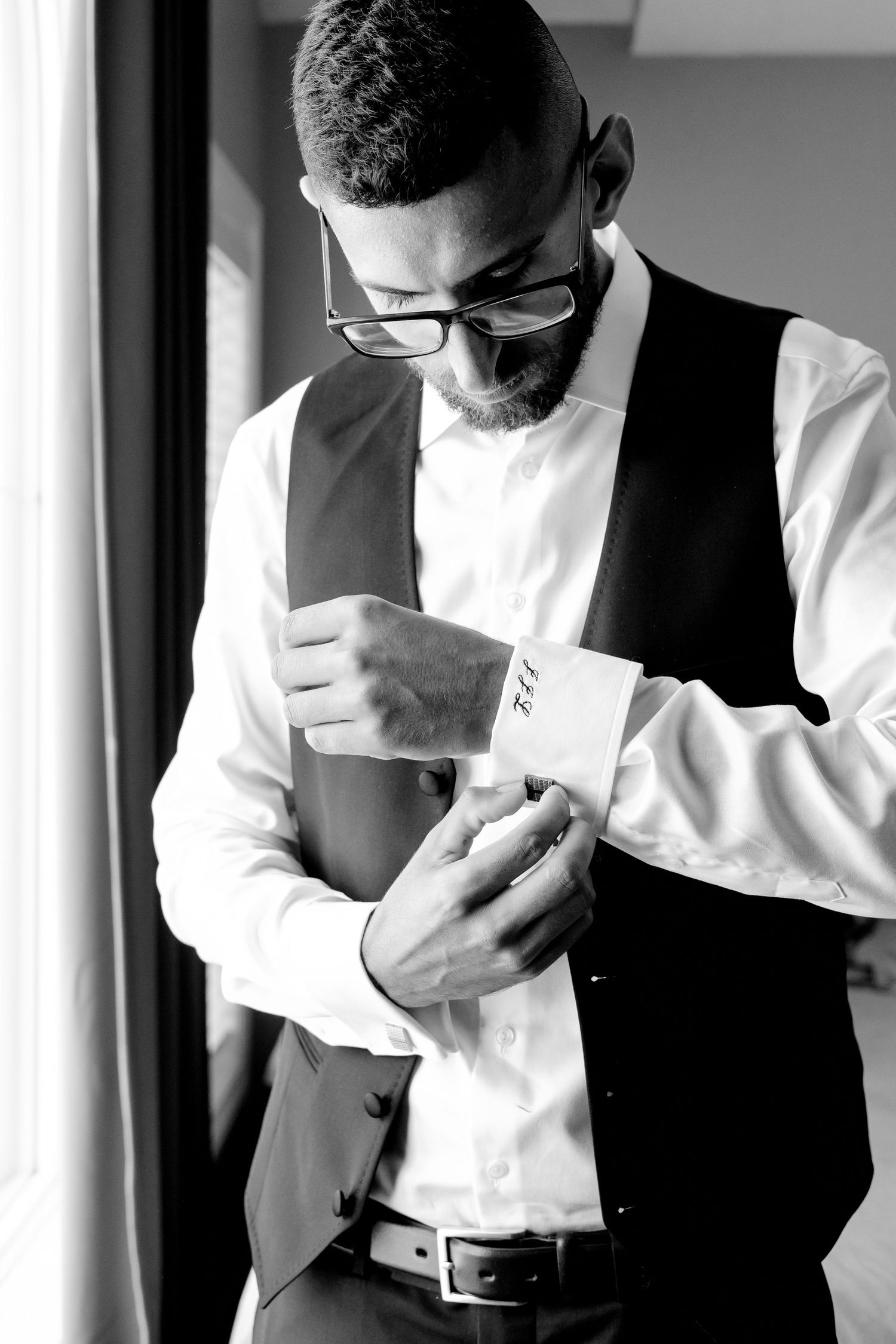 A person in a vest and white shirt adjusts their cufflinks in natural window light.