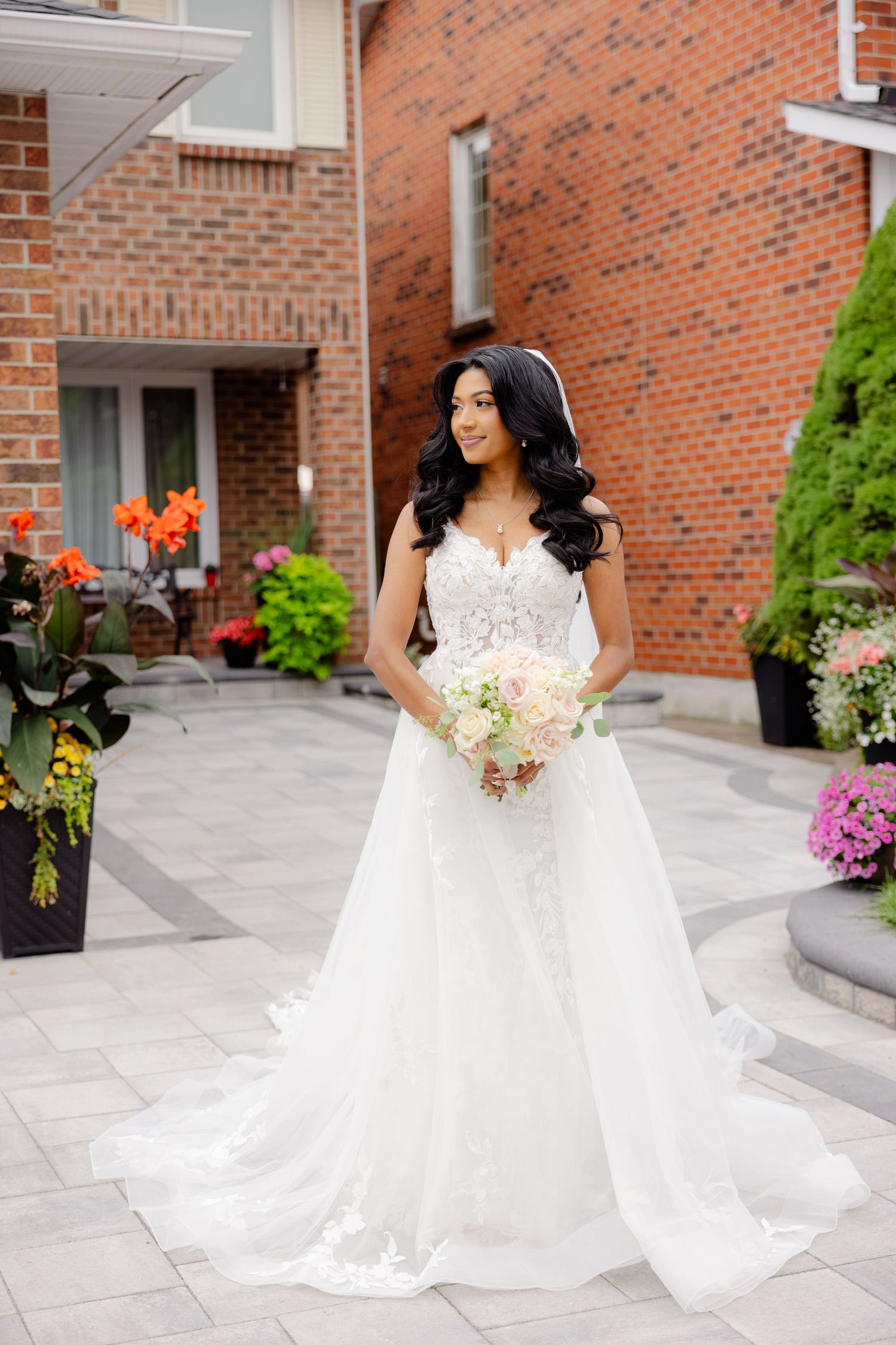 Elegant bride with cascading veil holds white bouquet against red brick exterior.