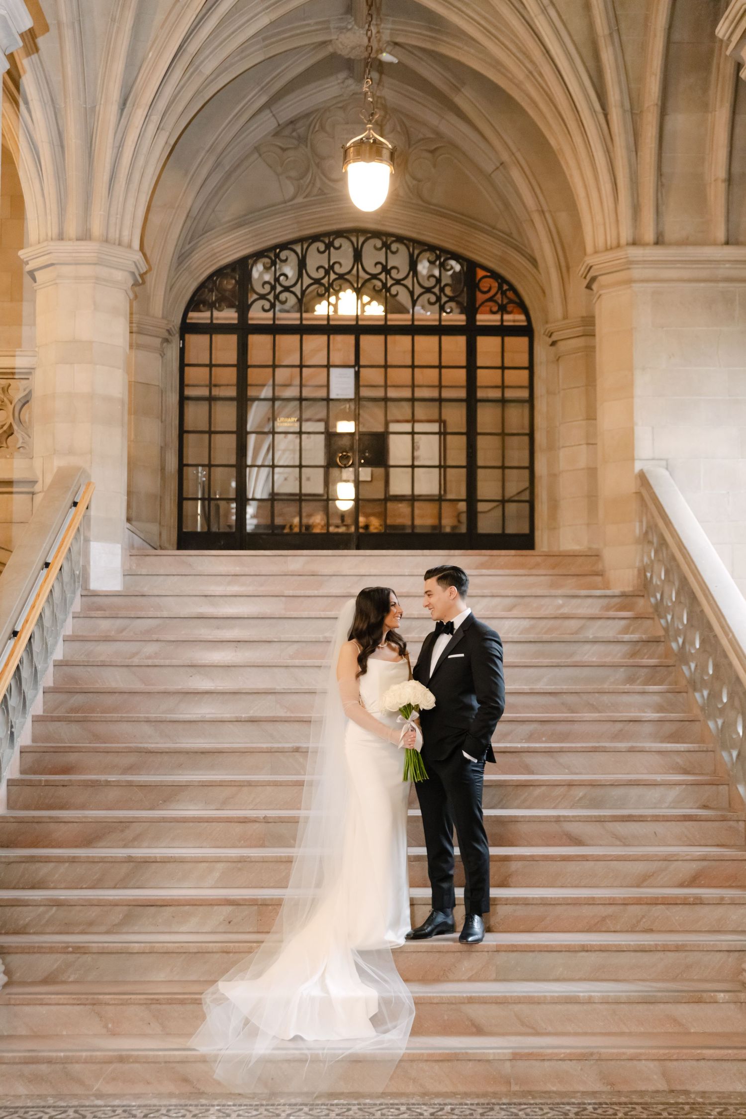 Wedding couple stands together on classic marble steps under vaulted ceiling and decorative window.