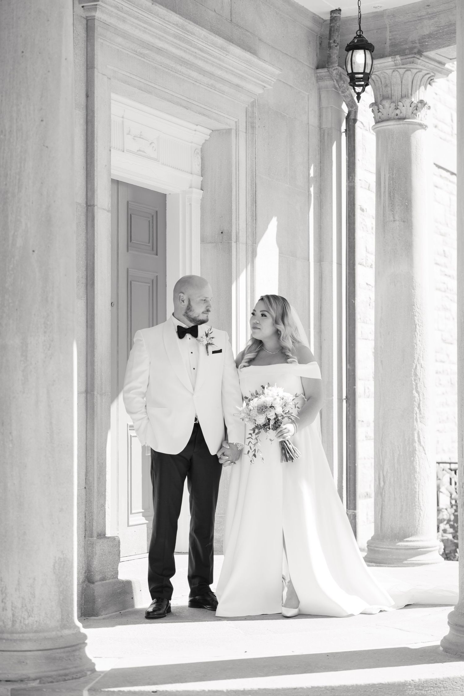 Black and white wedding portrait showing couple standing between classical columns with dramatic lighting.