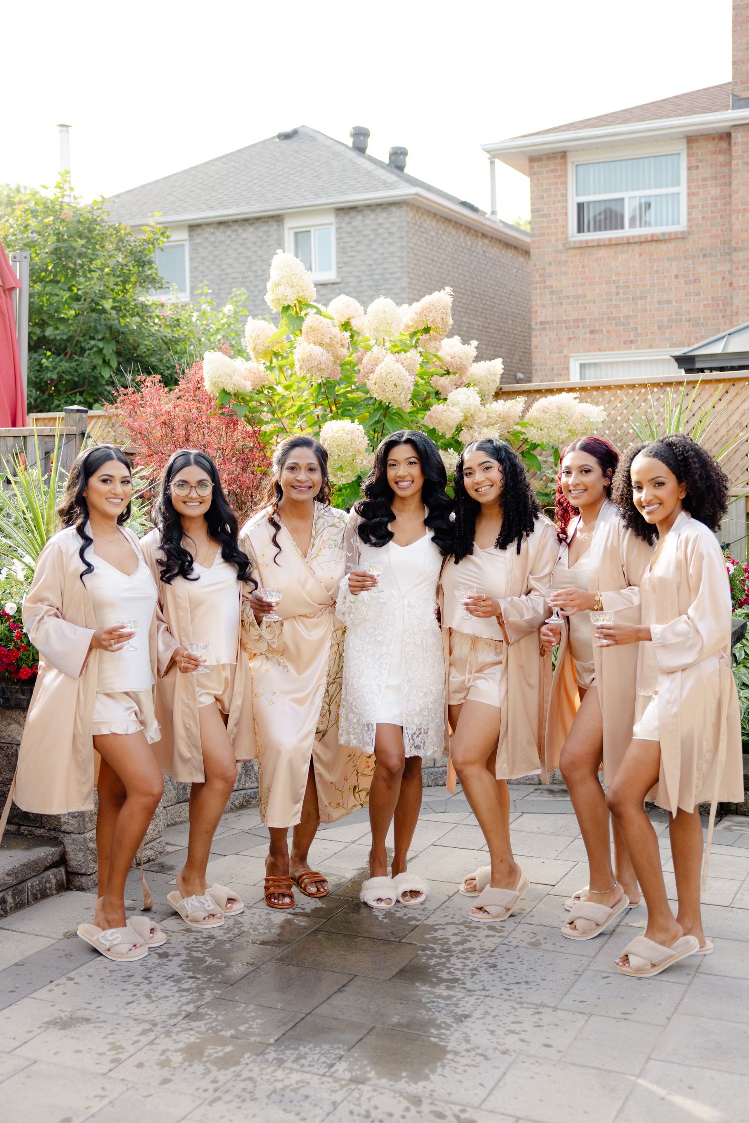 Bridal party poses in matching silk robes on a patio with beautiful floral landscaping.