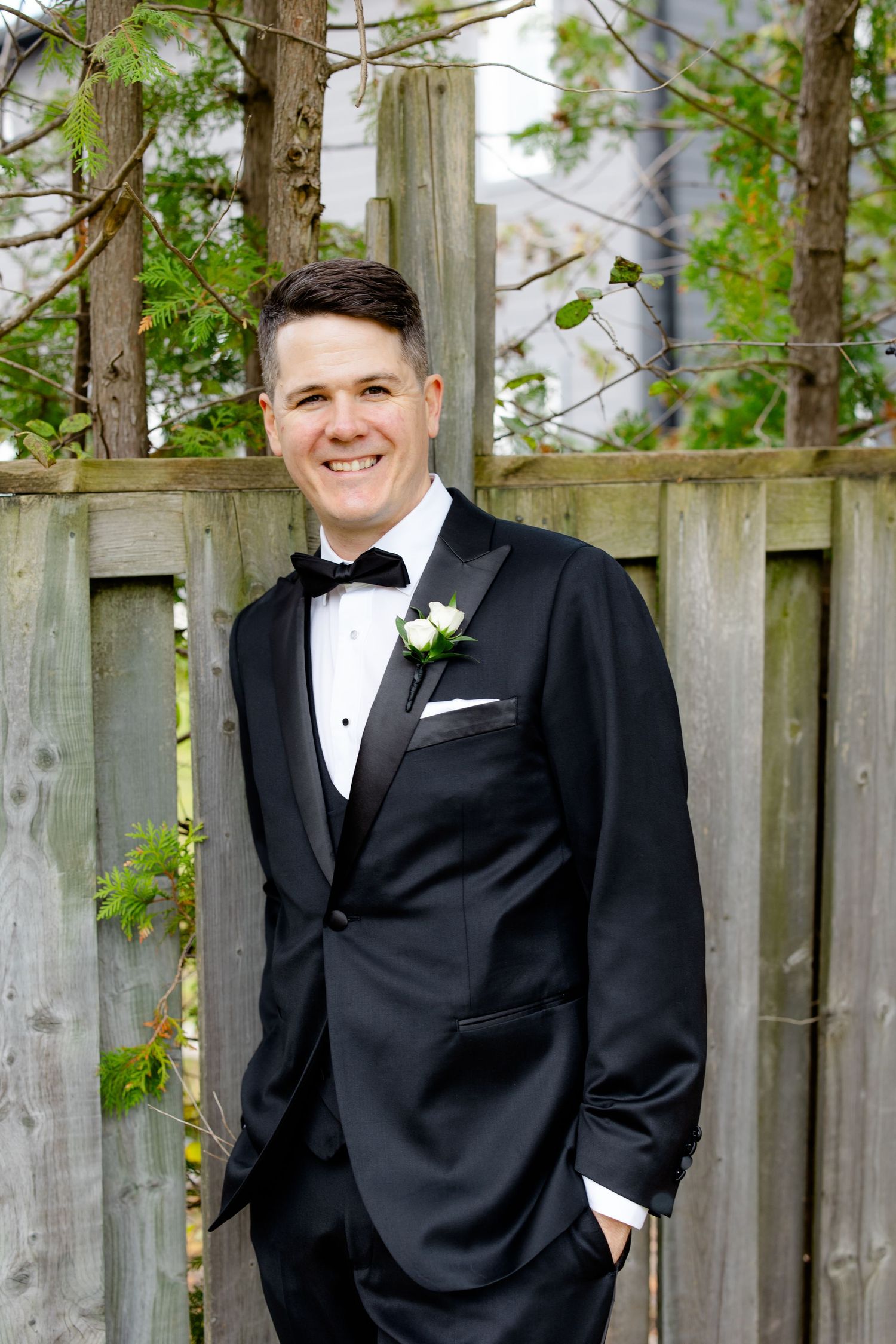 Formal portrait of a groom wearing a black tuxedo and white boutonniere against a wooden fence backdrop.