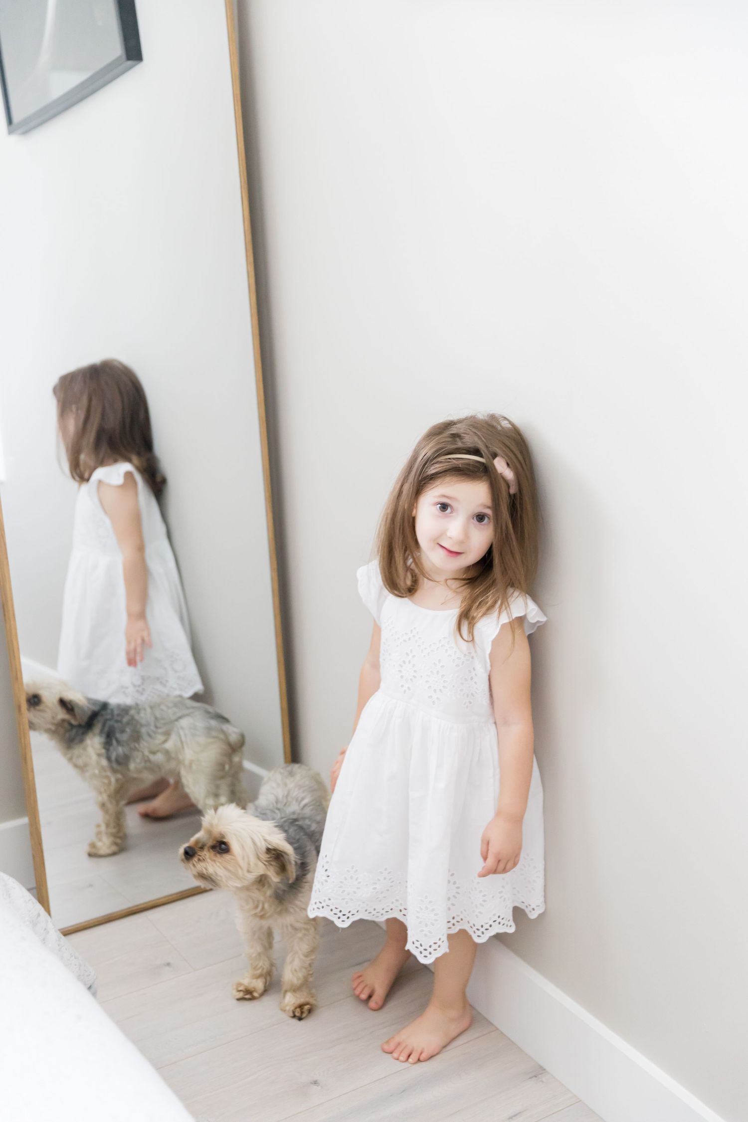 Little girl in white dress stands beside full-length mirror with small dog.