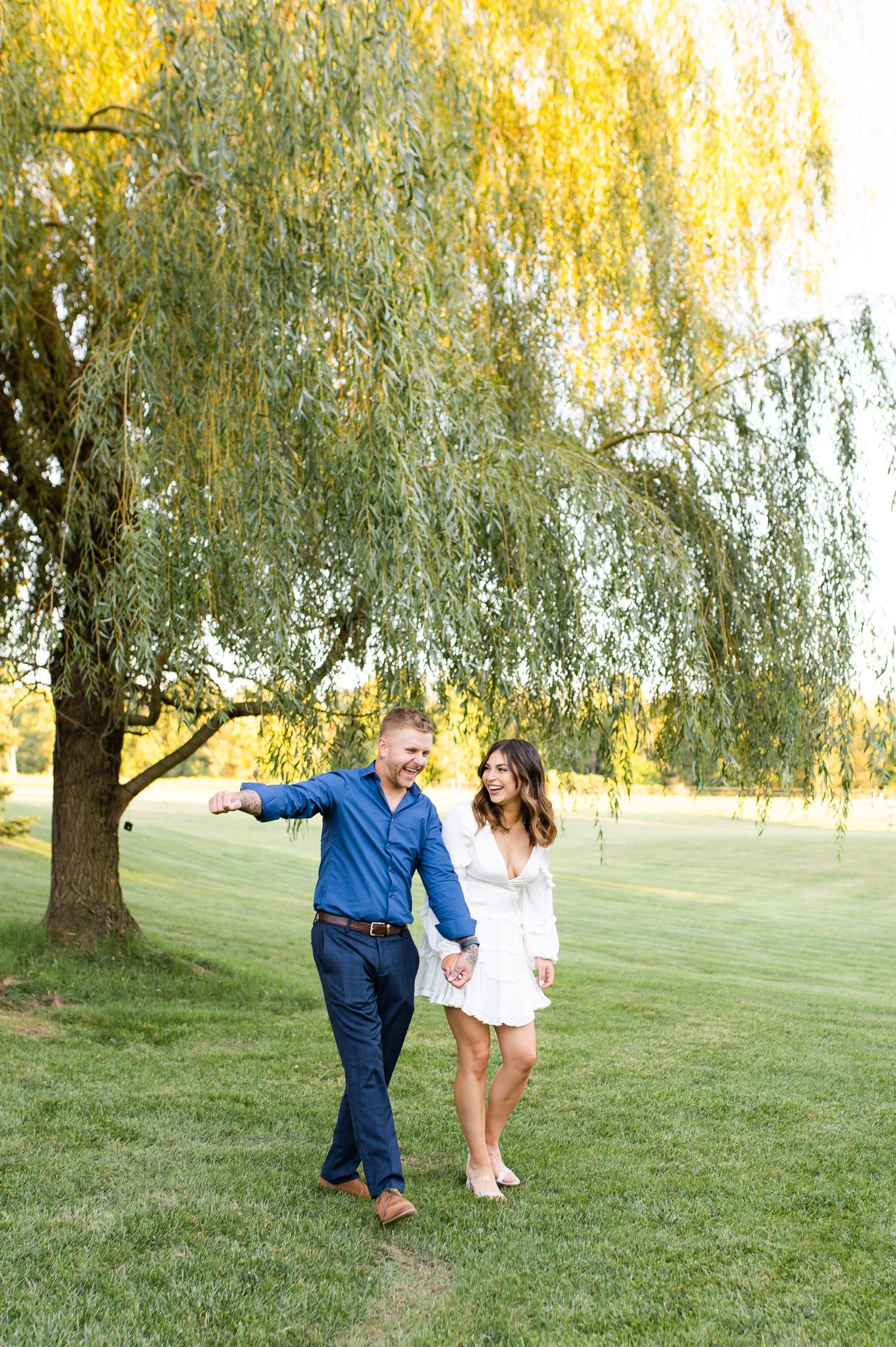 A romantic couple walks together on grassy lawn beneath a willow tree during golden hour.