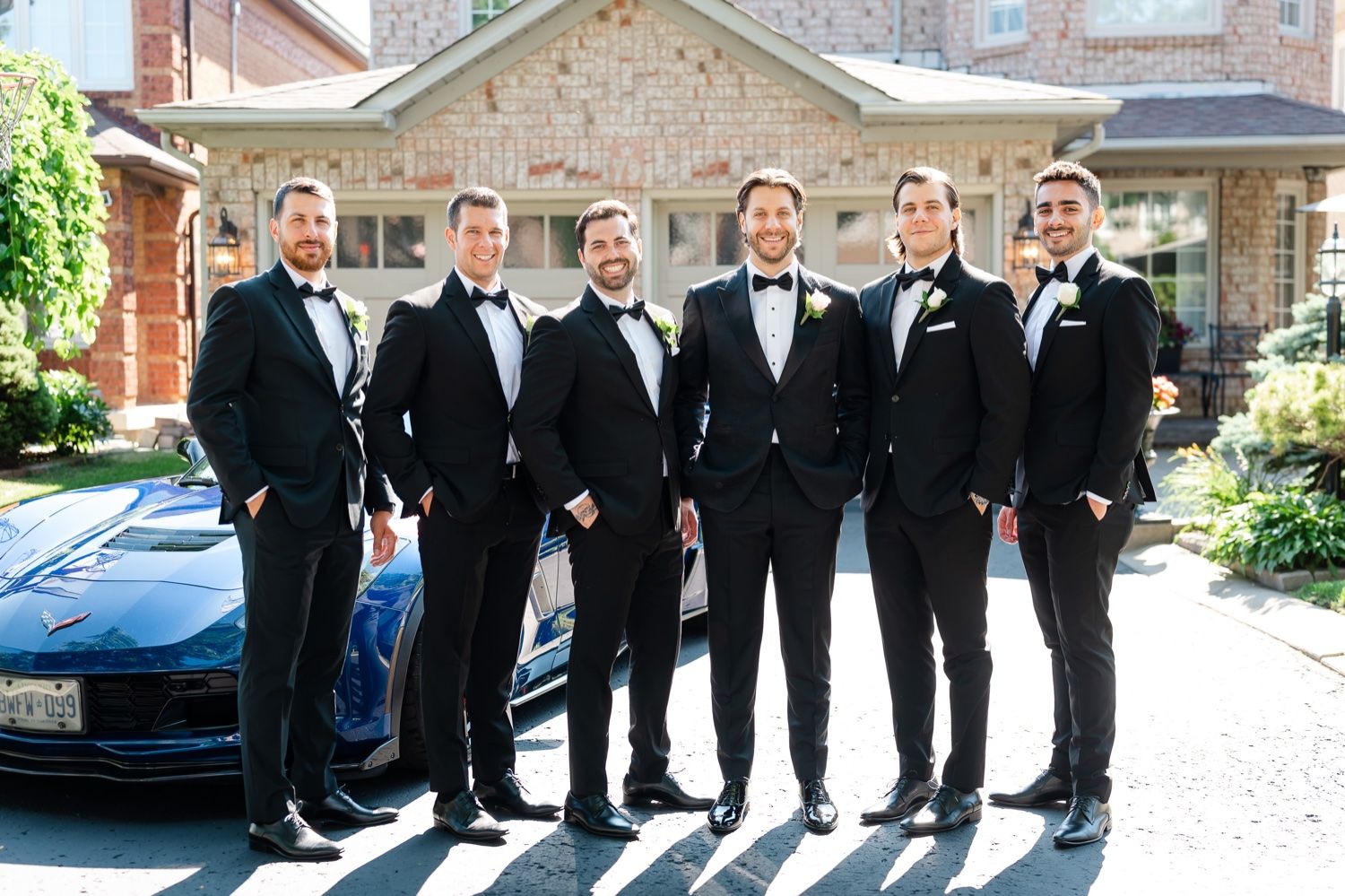 Groomsmen in black tuxedos pose together next to blue sports car.
