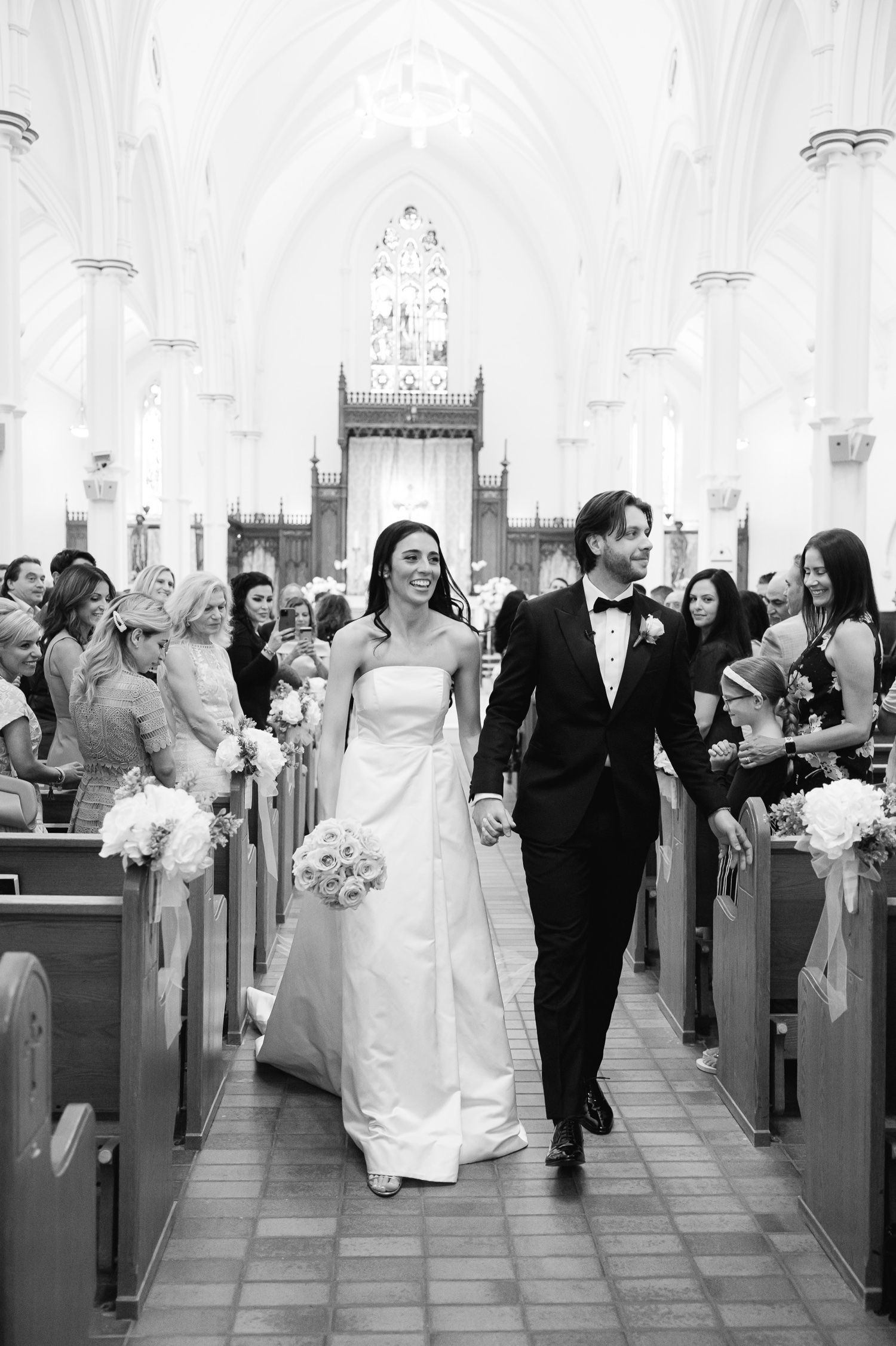 A couple processes down a church aisle while guests watch from wooden pews.