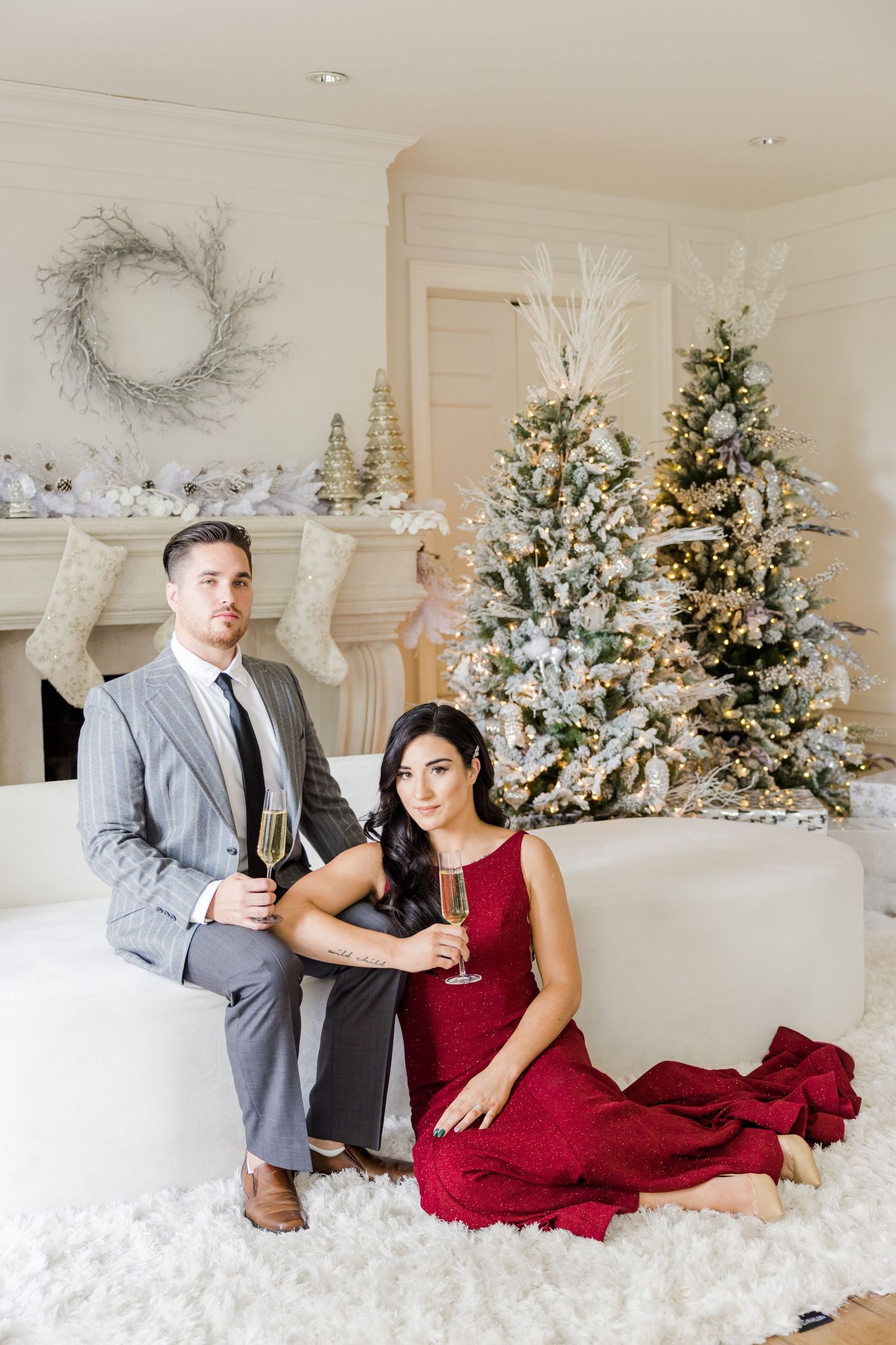 Couple in formal wear posing on white couch with flocked Christmas trees in background.
