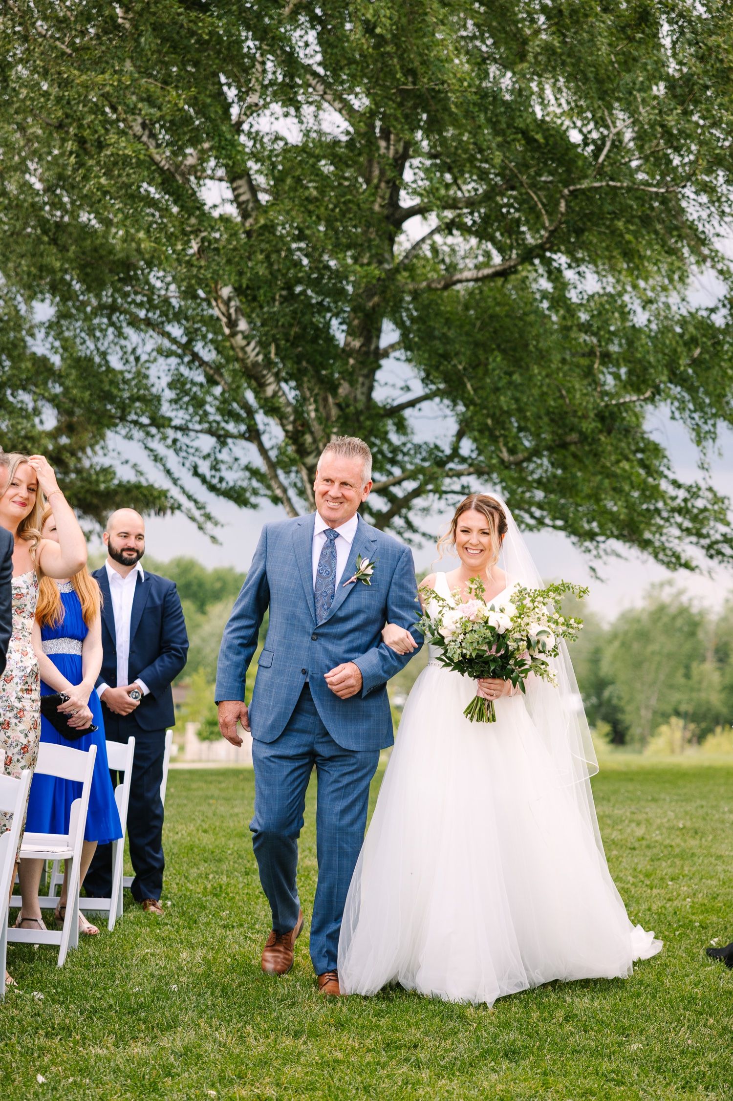 Wedding processional on grassy lawn under large birch tree with guests watching.