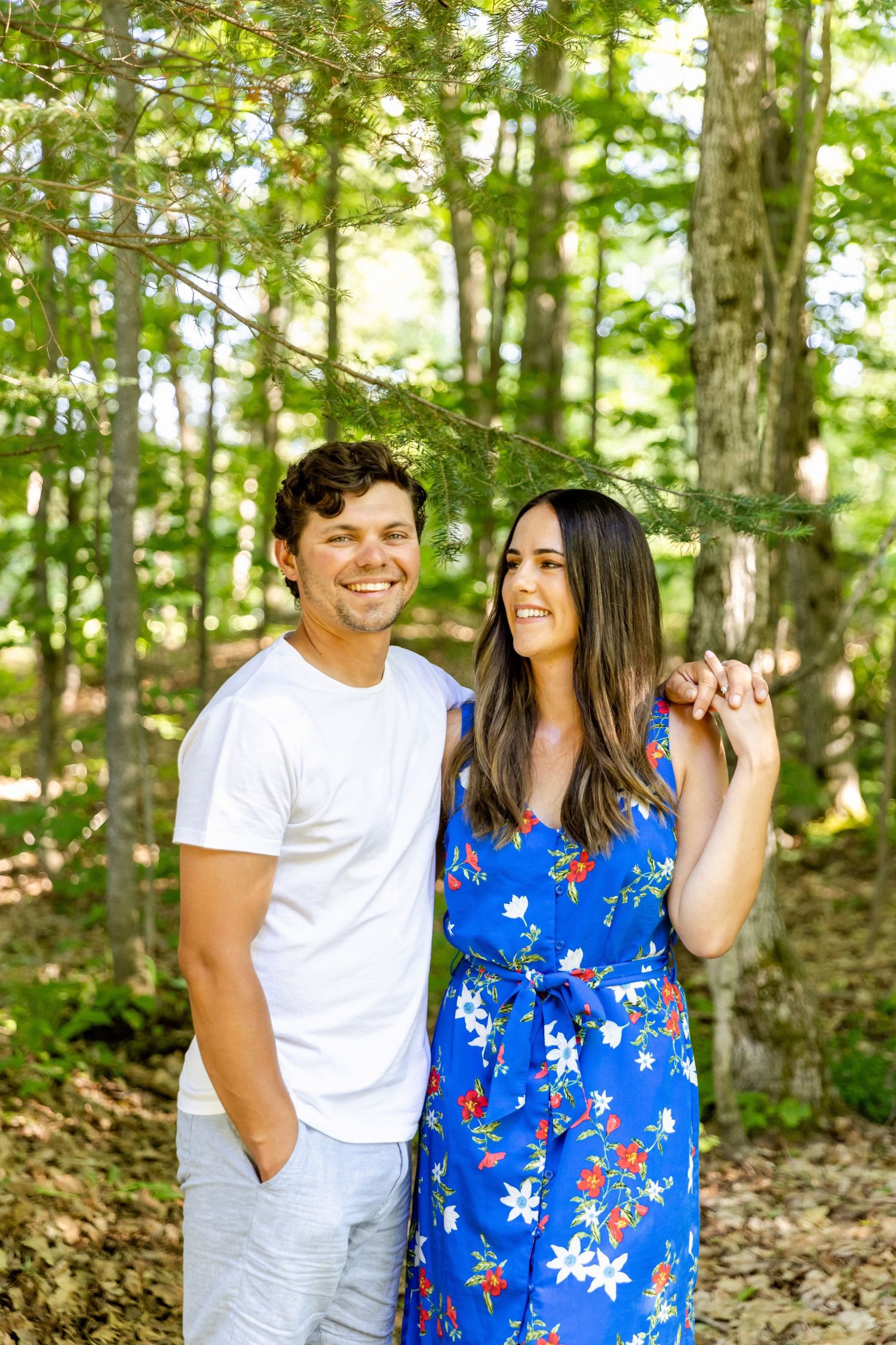 Couple in summer attire smiling together against a lush forest backdrop.