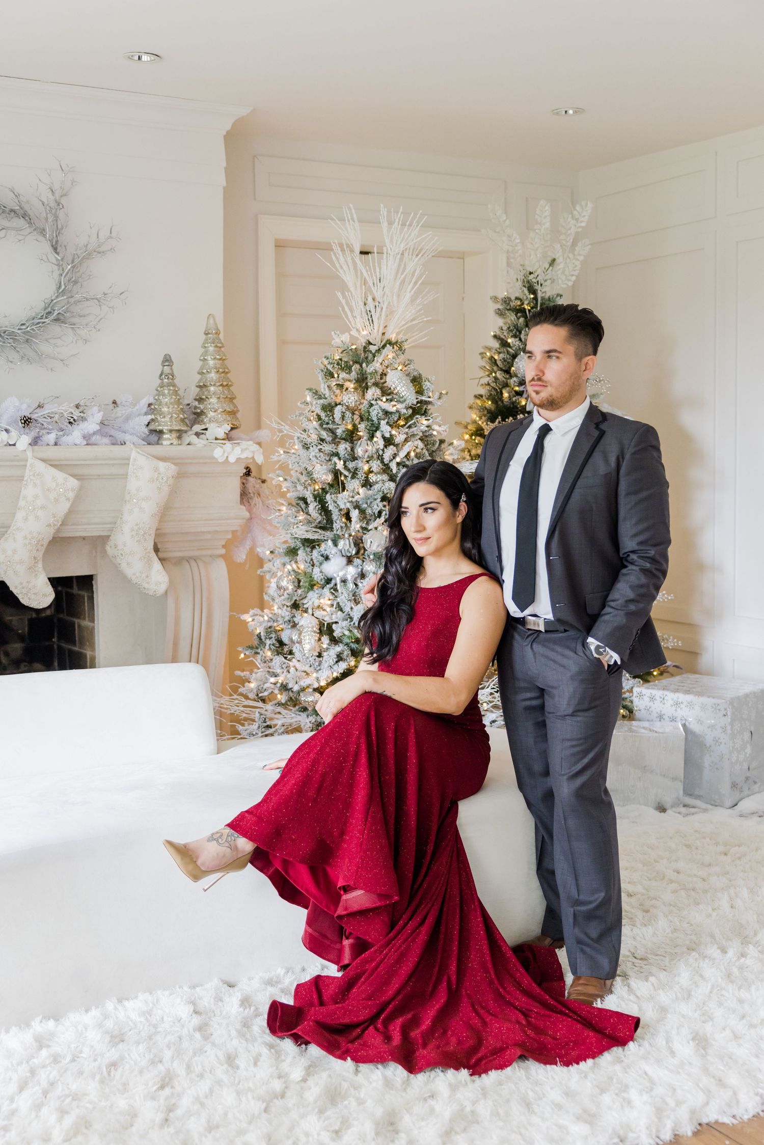 Elegant couple poses by Christmas trees with white decorations and stockings in a festive holiday setting.