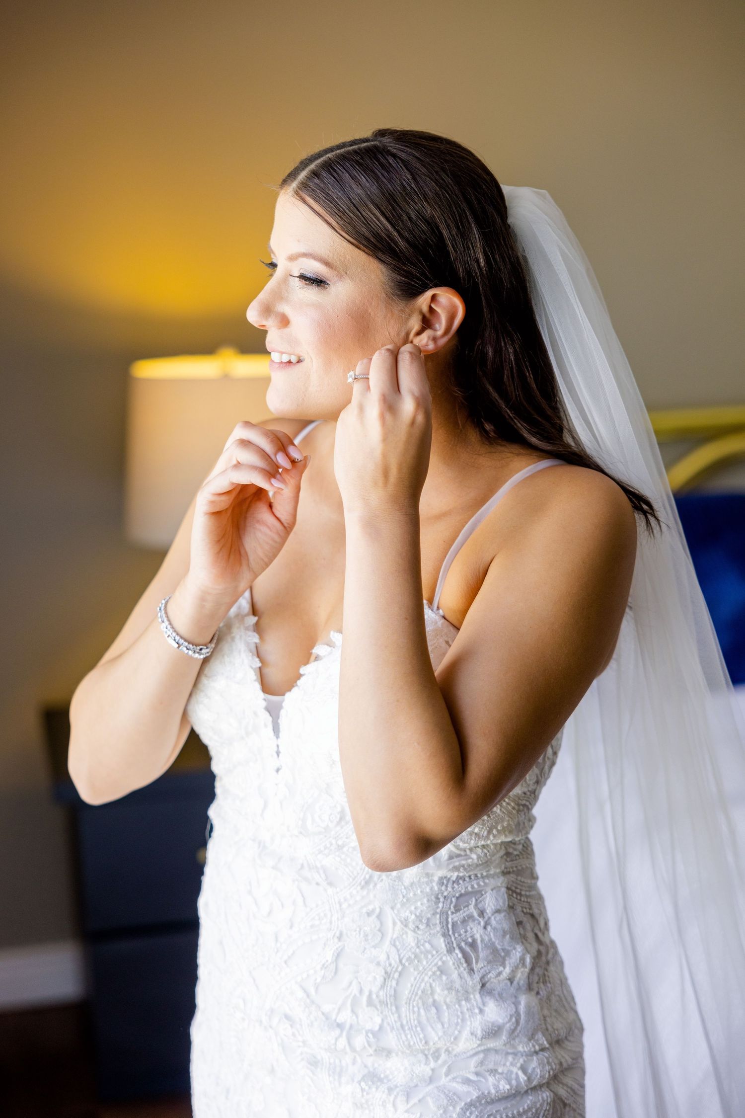 Bride in lace dress and veil adjusting earrings with warm lighting in background.